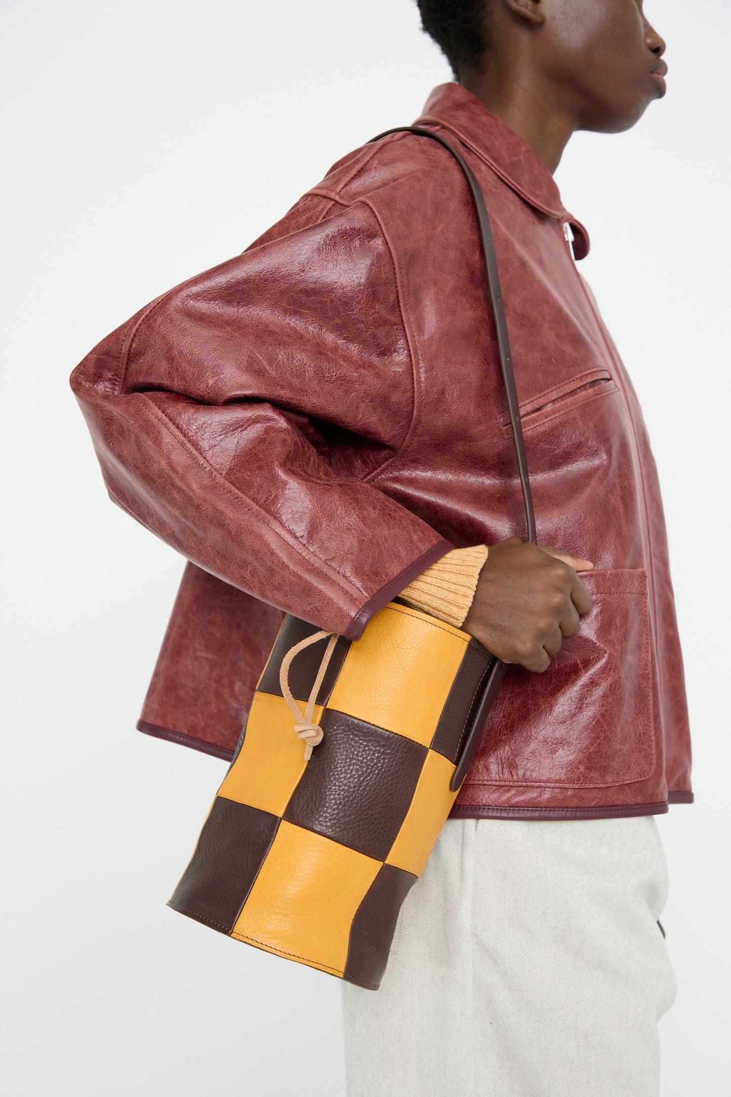 A person wearing a brown leather jacket carries the Cawley Leather Patchwork Bucket Bag in Tan and Mustard, featuring a chic checkered design with an adjustable strap.