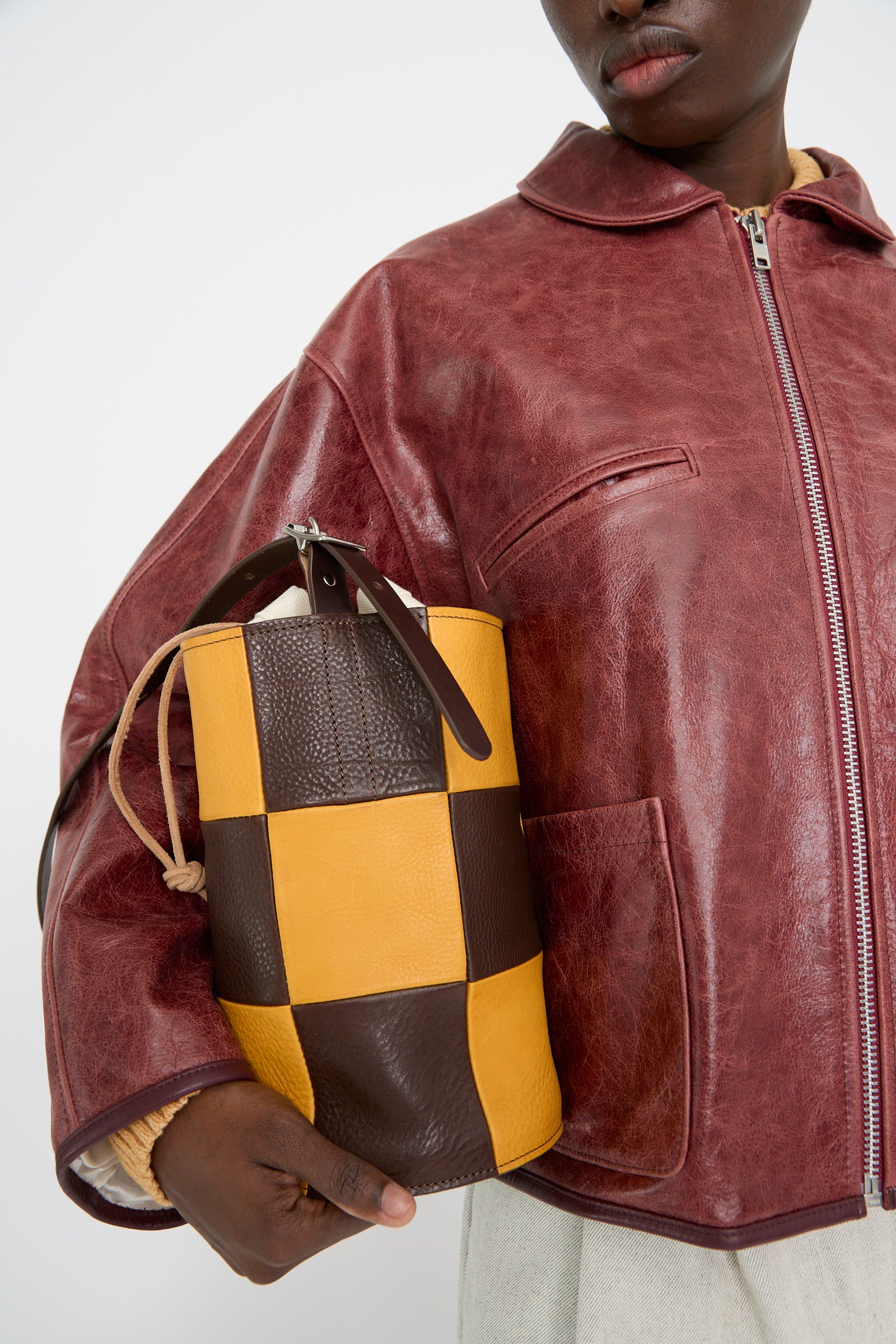 A person wearing a maroon leather jacket holds the Cawley Leather Patchwork Bucket Bag in Tan and Mustard, featuring an adjustable strap for convenience.