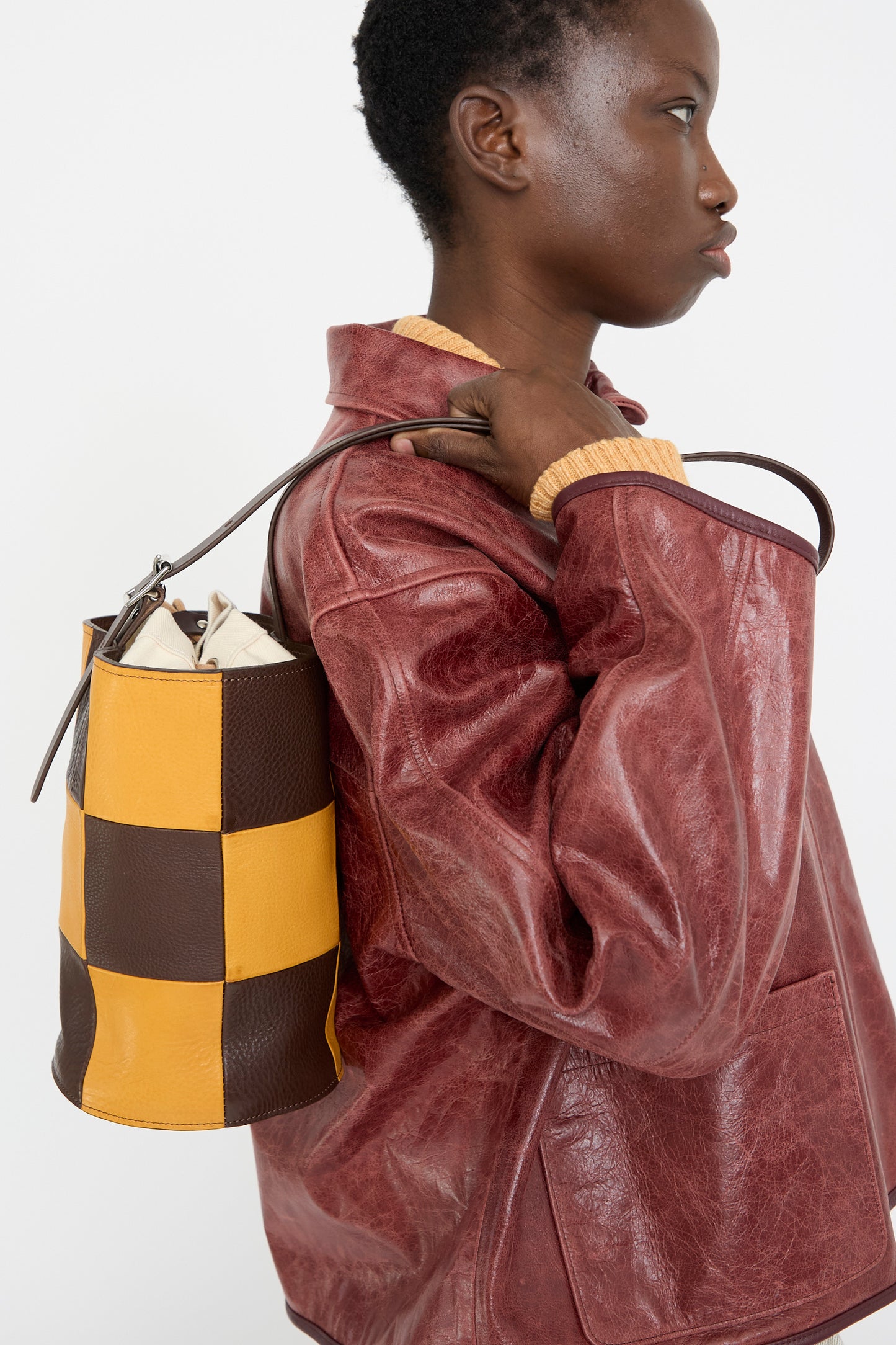 A person in a red leather jacket holds Cawley's Leather Patchwork Bucket Bag in Tan and Mustard, featuring a stylish checkered design and adjustable shoulder strap.