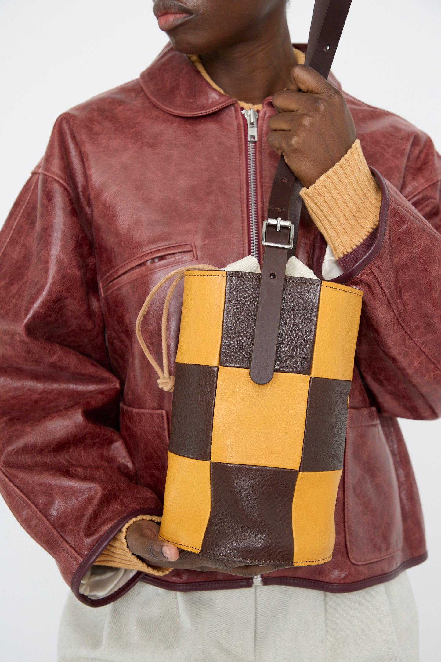 A person in a red leather jacket holds a Cawley Leather Patchwork Bucket Bag in Tan and Mustard, featuring an adjustable shoulder strap and crafted from grainy patchwork leather.