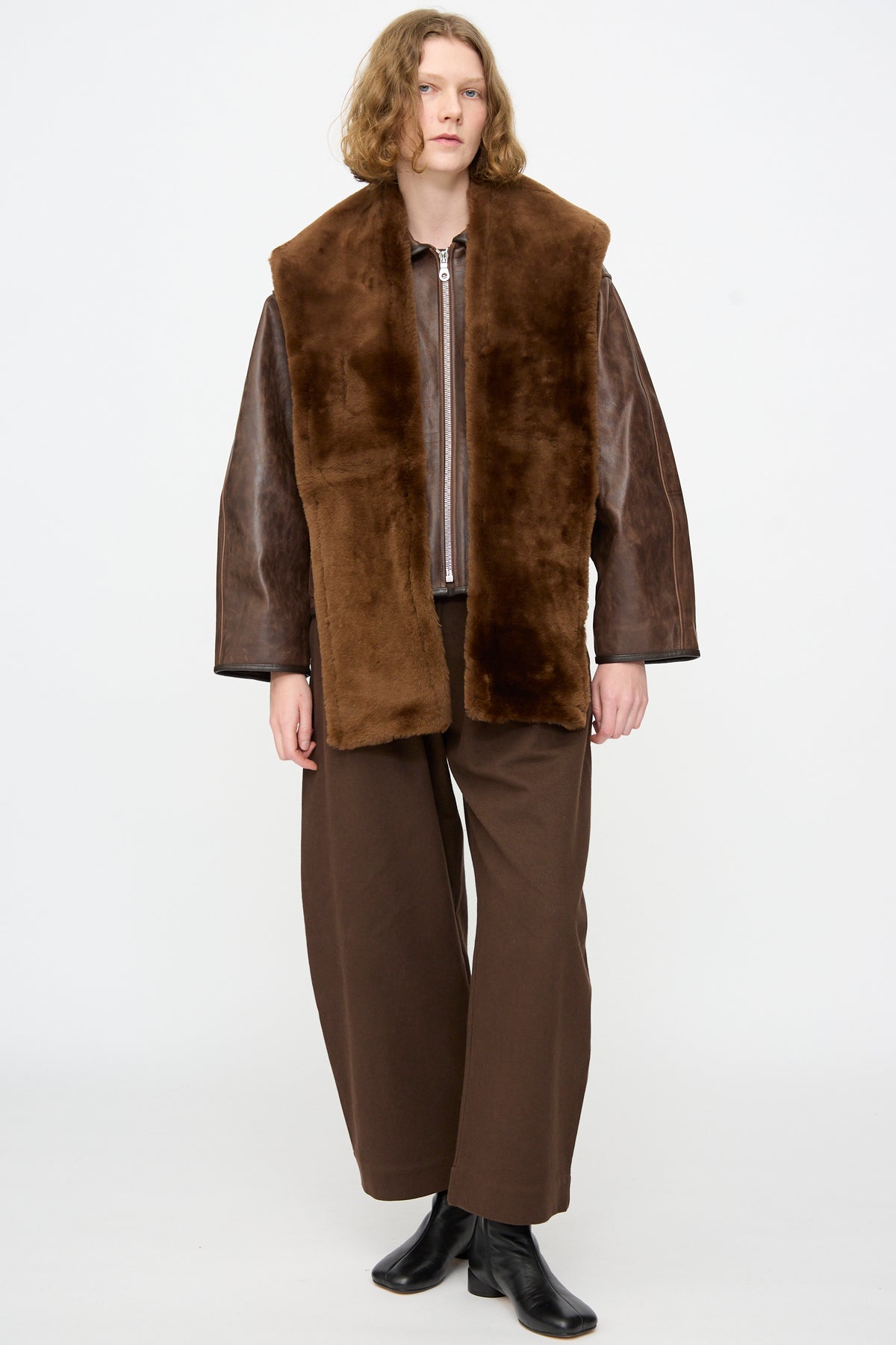 A person wears Cawley’s Leather Sheepskin Stole in Walnut draped over a brown leather jacket, paired with wide brown pants and black boots, standing against a plain white background.