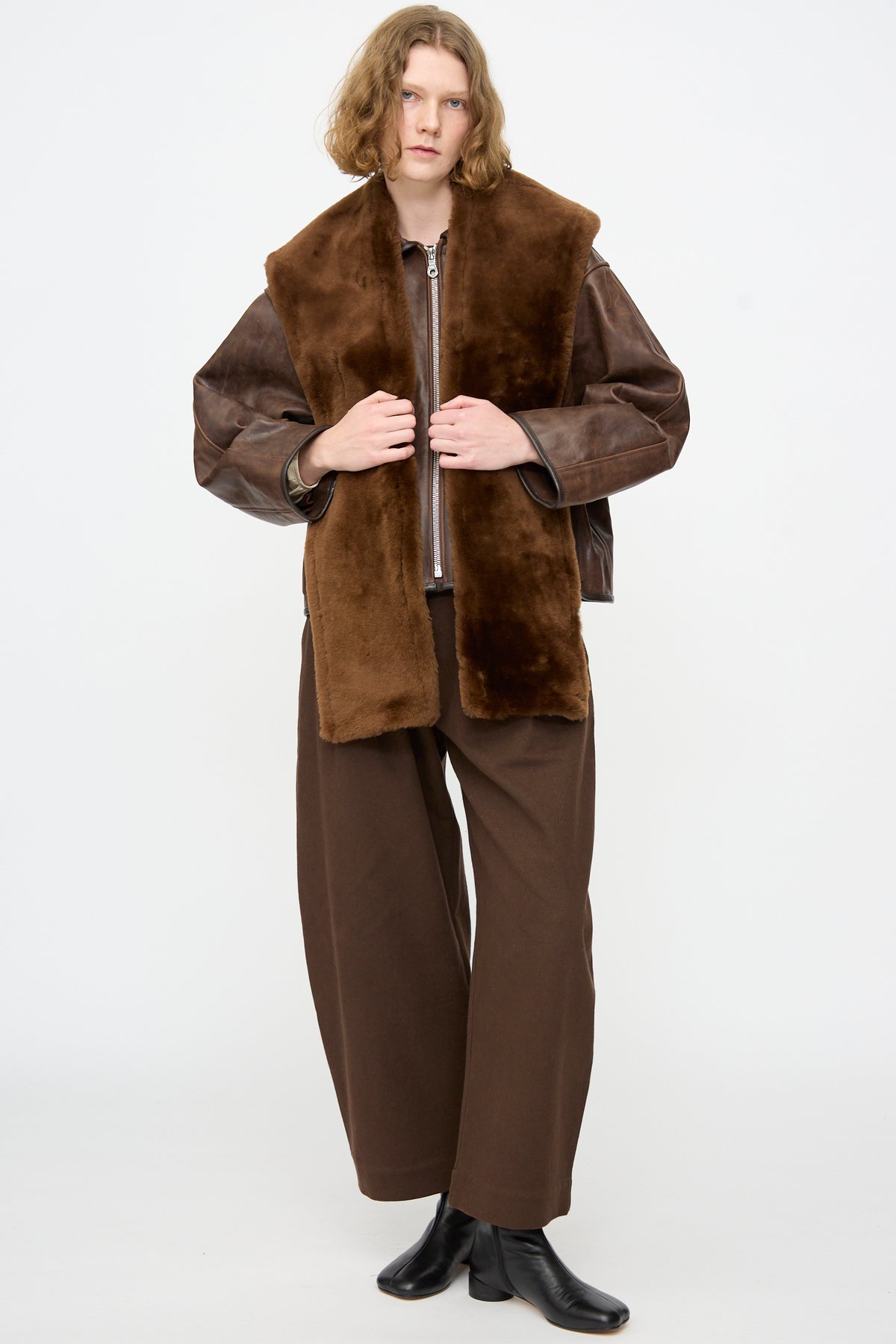 A person stands against a plain background wearing the Cawley Leather Sheepskin Stole in Walnut, styled with brown wide-leg pants, a brown jacket, and black boots.