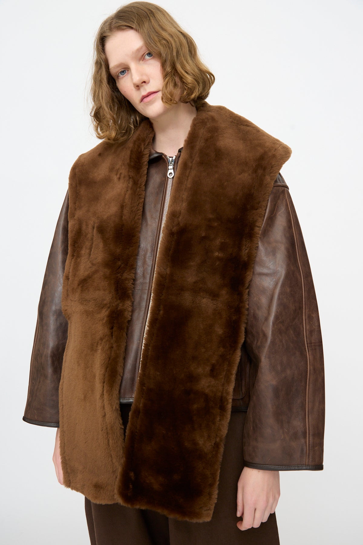A person with shoulder-length hair wears a brown leather jacket and the Cawley Leather Sheepskin Stole in Walnut against a plain background.