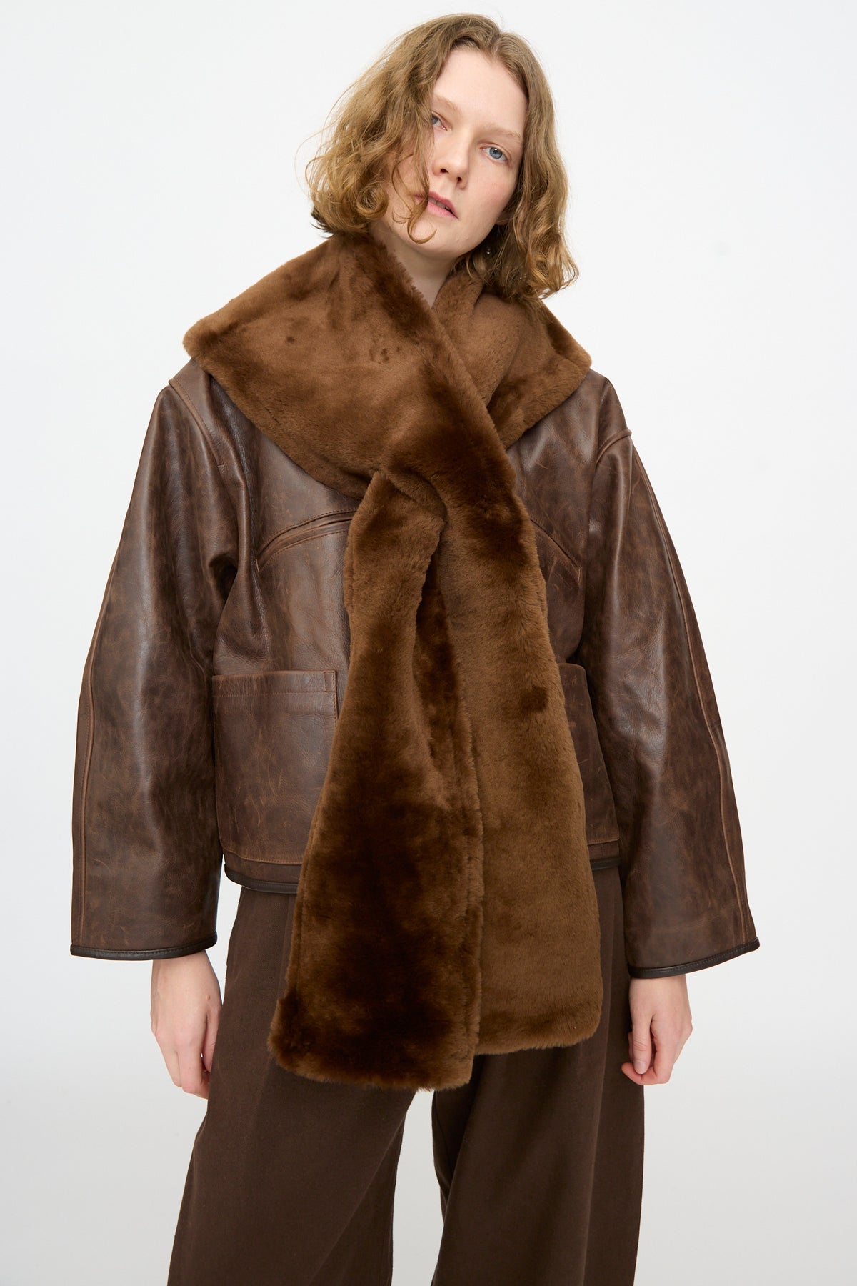Model is wearing the Leather Sheepskin Stole in Walnut by Cawley, paired with a brown leather jacket and wide brown pants, standing against a plain white background.