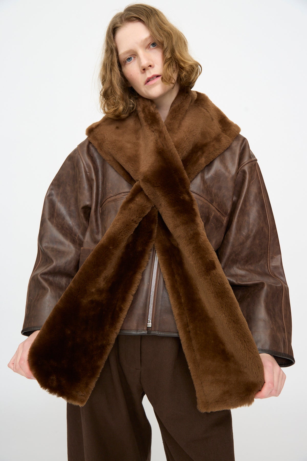 A model wears the Cawley Leather Sheepskin Stole in Walnut, crossed at the front over a brown leather jacket, set against a plain white background.