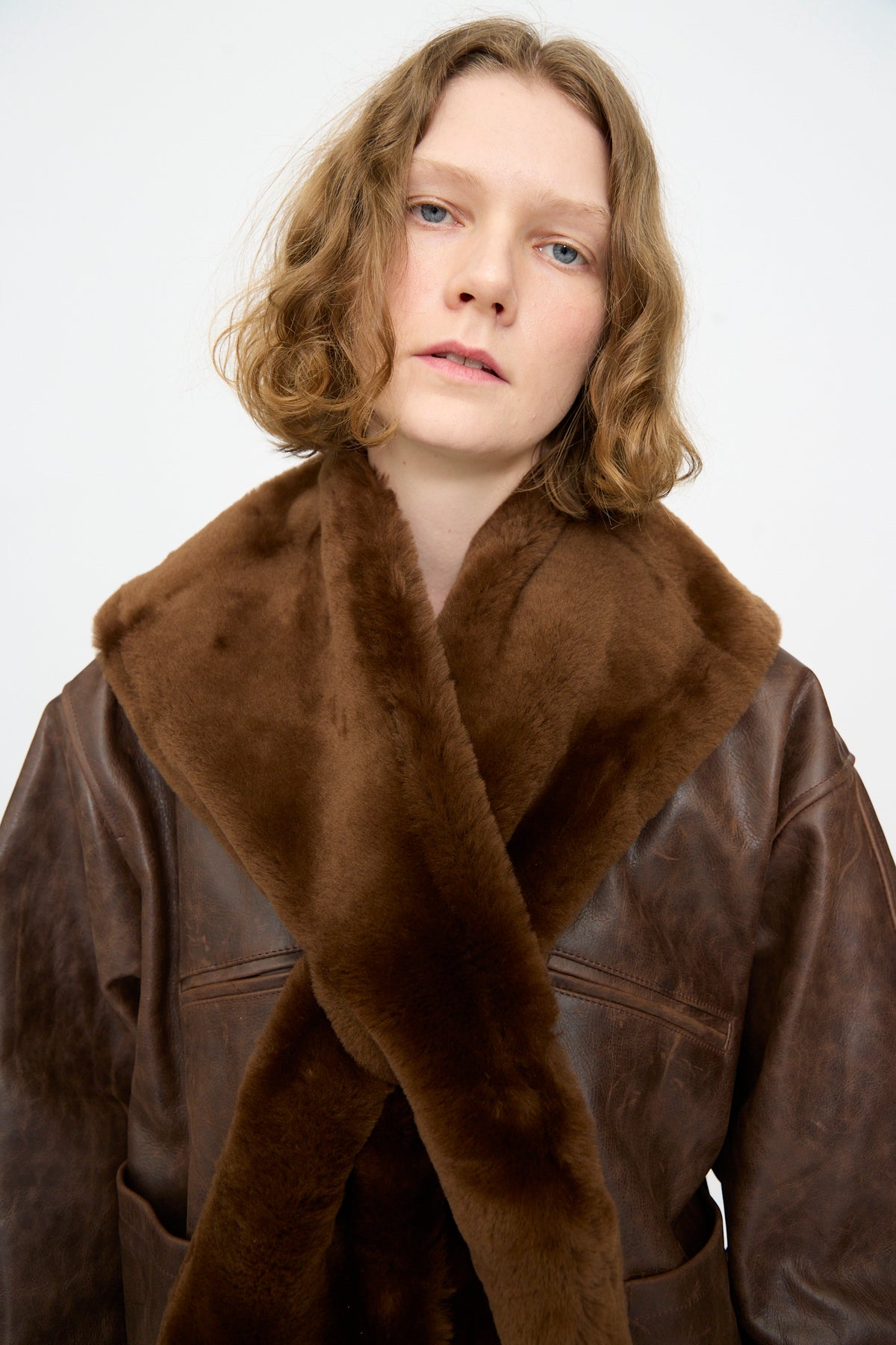 A person with wavy, shoulder-length hair wears a brown leather coat featuring the Cawley Leather Sheepskin Stole in Walnut, which has a large, plush walnut collar with leather backing, standing against a plain white background.