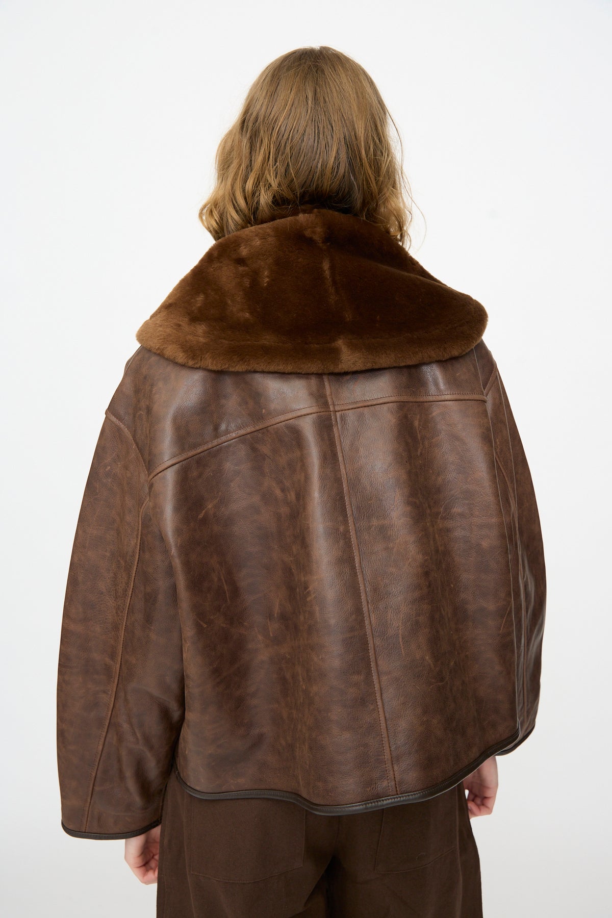 A person with wavy light brown hair is seen from the back, wearing the Cawley Leather Sheepskin Stole in Walnut—a brown leather jacket featuring a wide, furry sheepskin stole collar—against a plain white background.