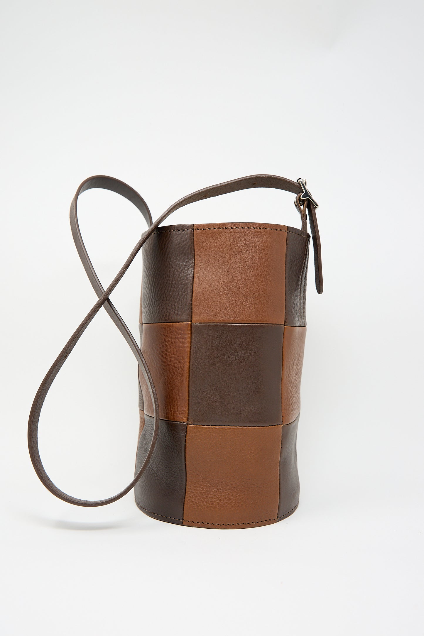 The Leather Bucket Bag in Chocolate and Tan by Cawley, a cylindrical Italian leather accessory adorned with a checkered patchwork pattern of brown and dark brown squares. It features a long adjustable strap against a white background.