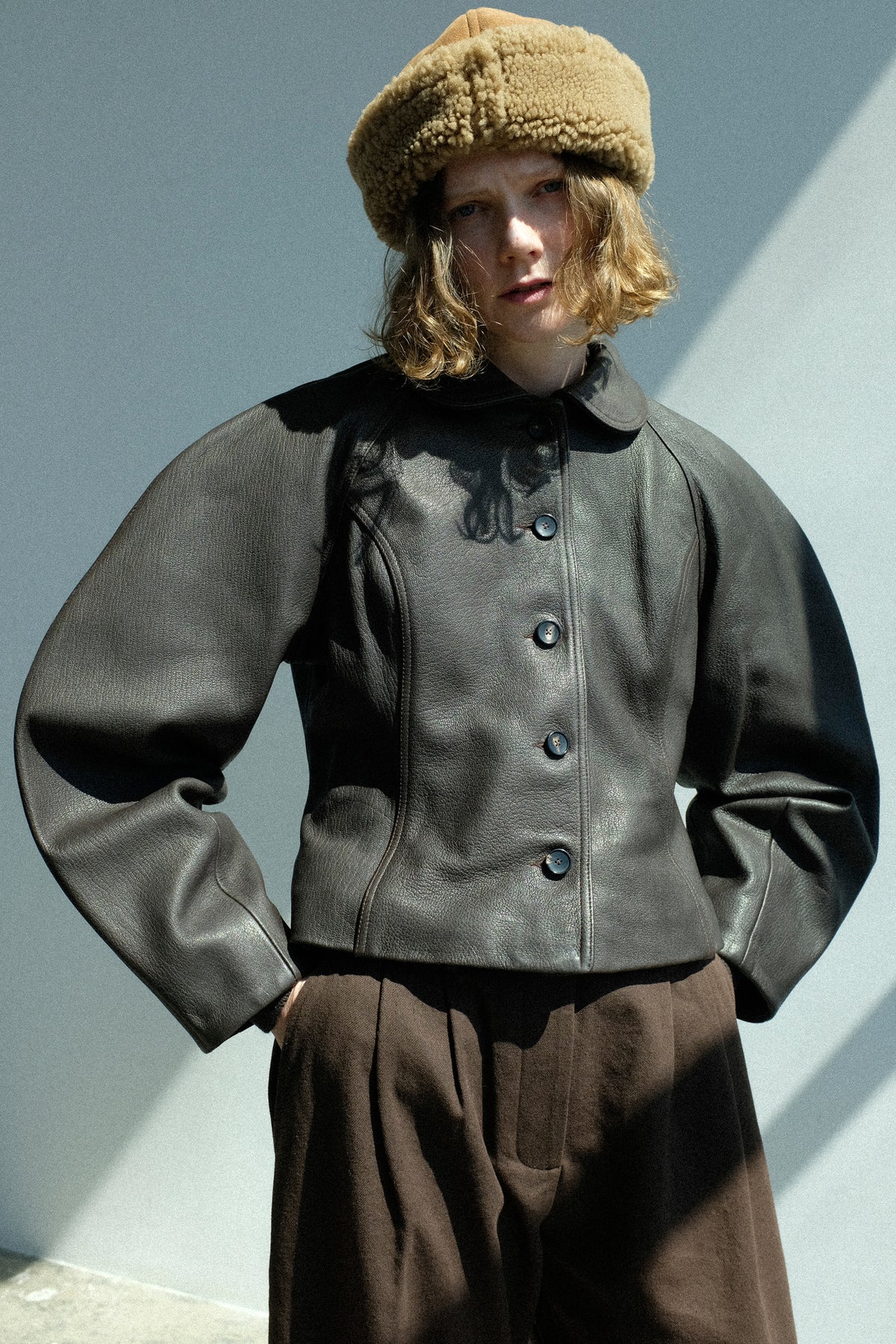 A model wearing Cawley's Leather Heidi Boned Jacket in Brown set against a minimalist gray wall with sharp shadows.