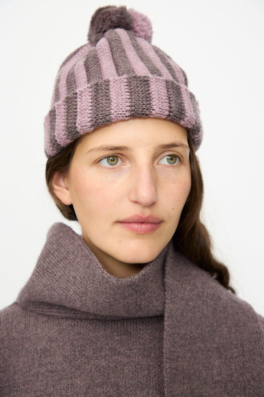 A person with light skin and long brown hair wears the Cawley Merino Wool Pleated Bobble Hat in Brezo and Luna, looking slightly to the right.