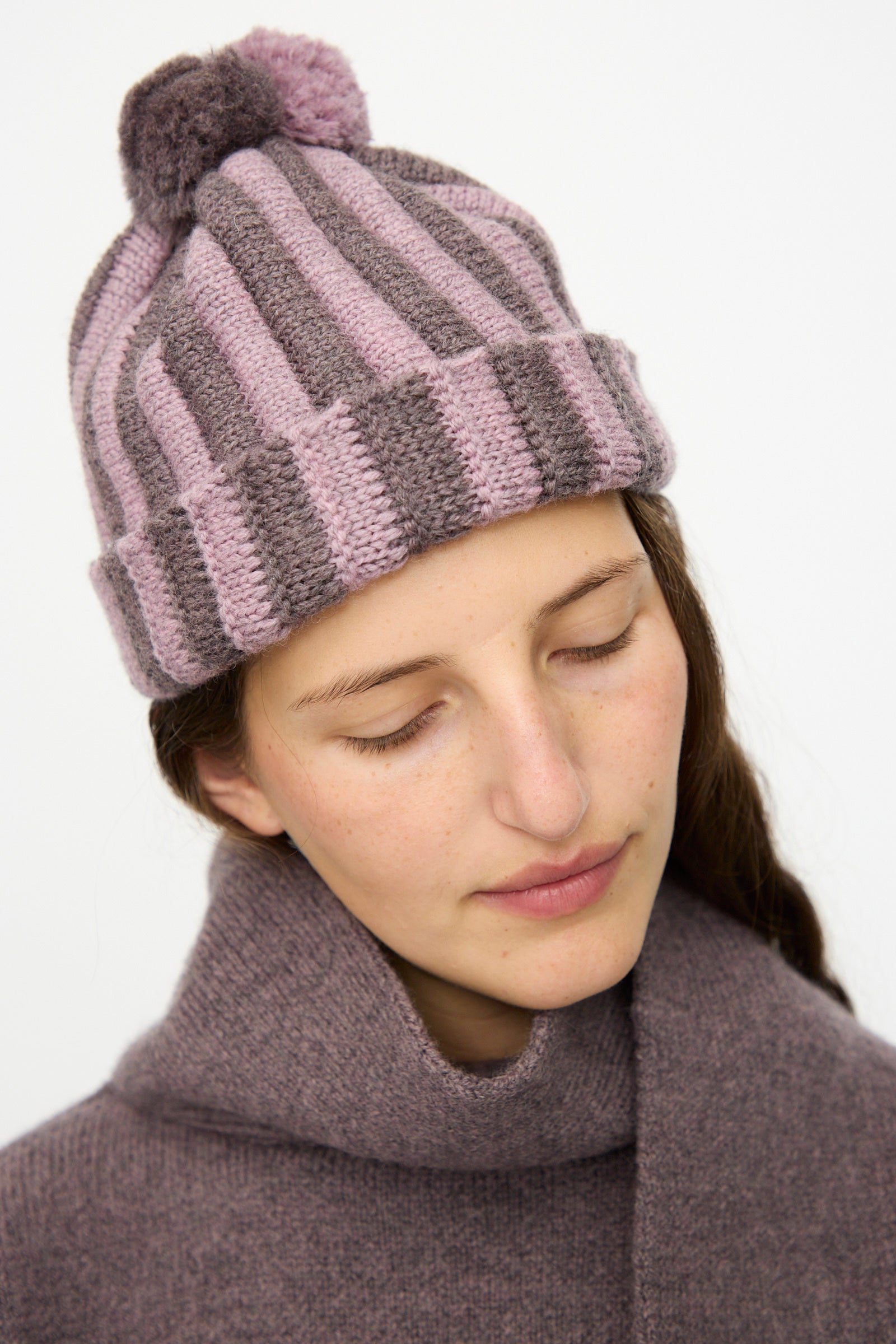 A woman in a Cawley Merino Wool Pleated Bobble Hat in Brezo and Luna gazes downward, her cozy knit accessories standing out against a plain background.
