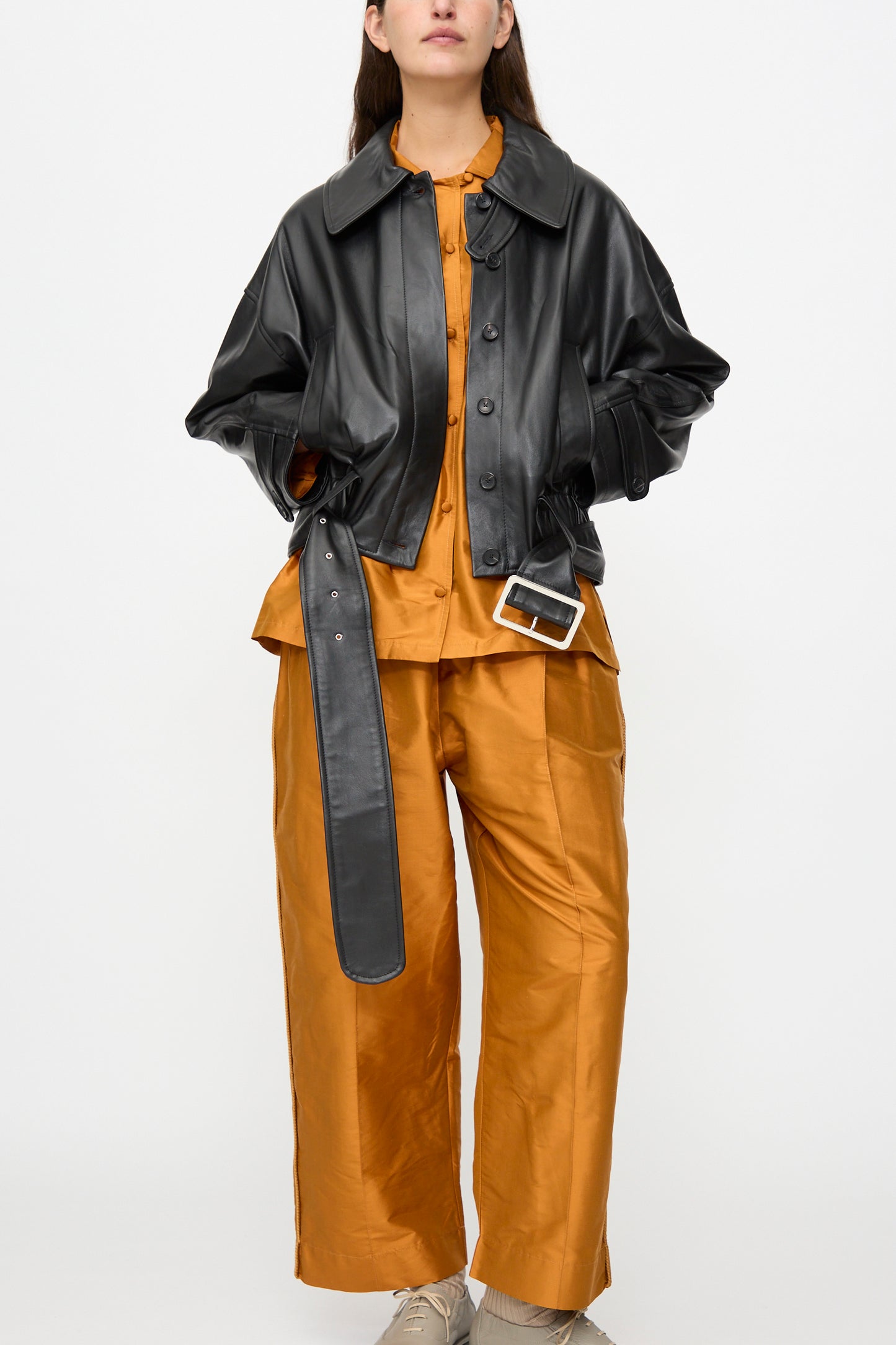 A woman wears the Cawley Nappa Leather Nara Jacket in Black, styled over an orange button-up shirt and matching wide-leg pants with beige shoes, standing against a plain white background.