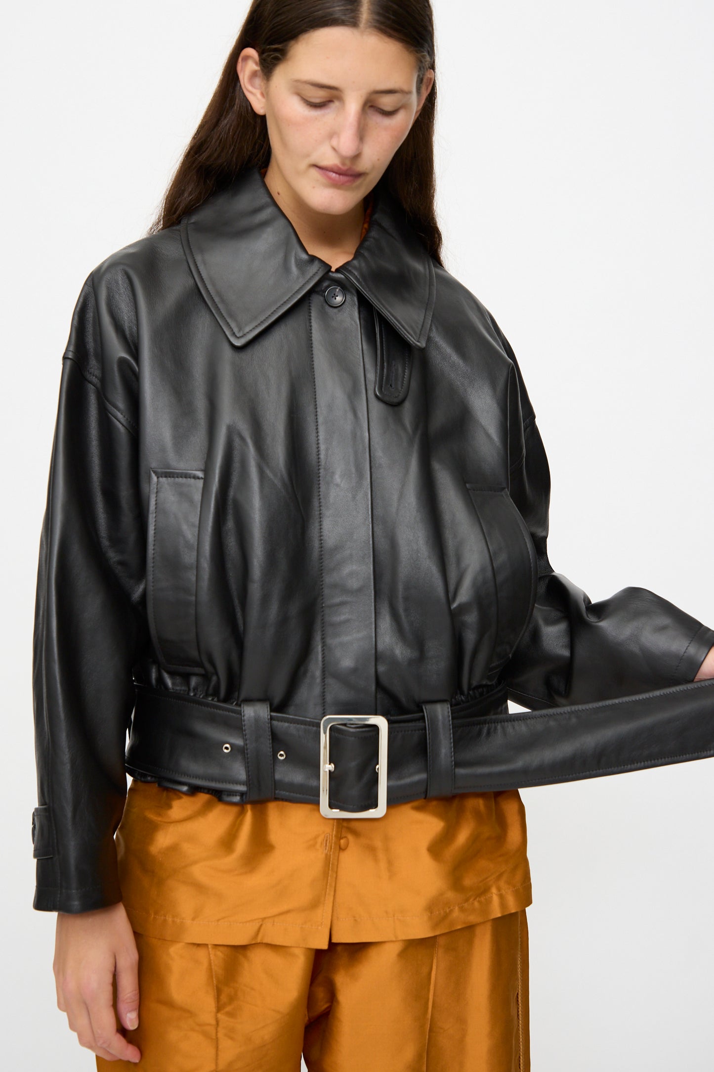 A person wears the Cawley Nappa Leather Nara Jacket in Black, styled with a wide belt and orange pants, looking down as they adjust the belt.