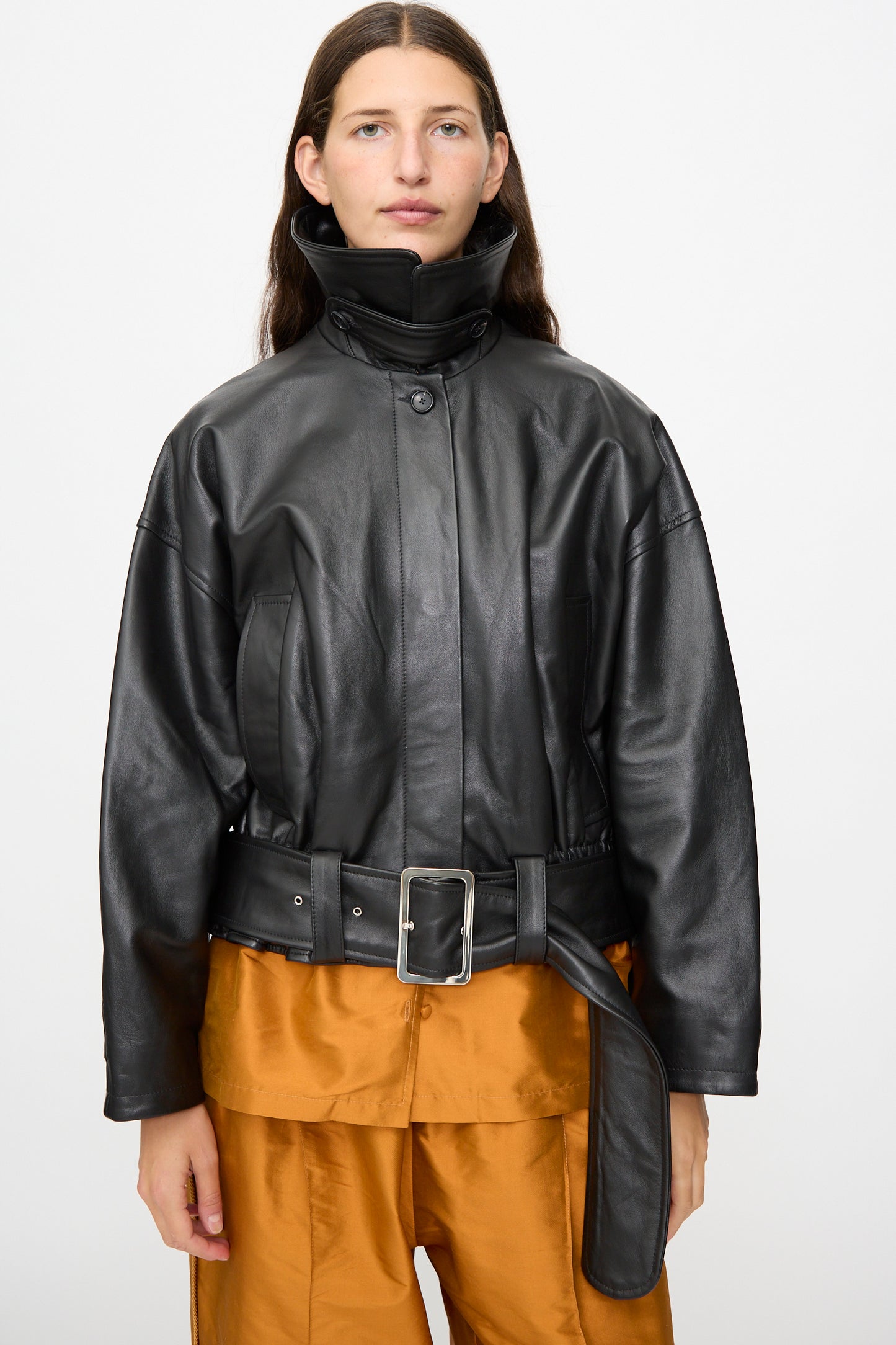 A woman wears the Cawley Nappa Leather Nara Jacket in Black—an oversized blouson with a high collar and belt—styled over an orange shirt and matching orange pants, standing against a plain white background.