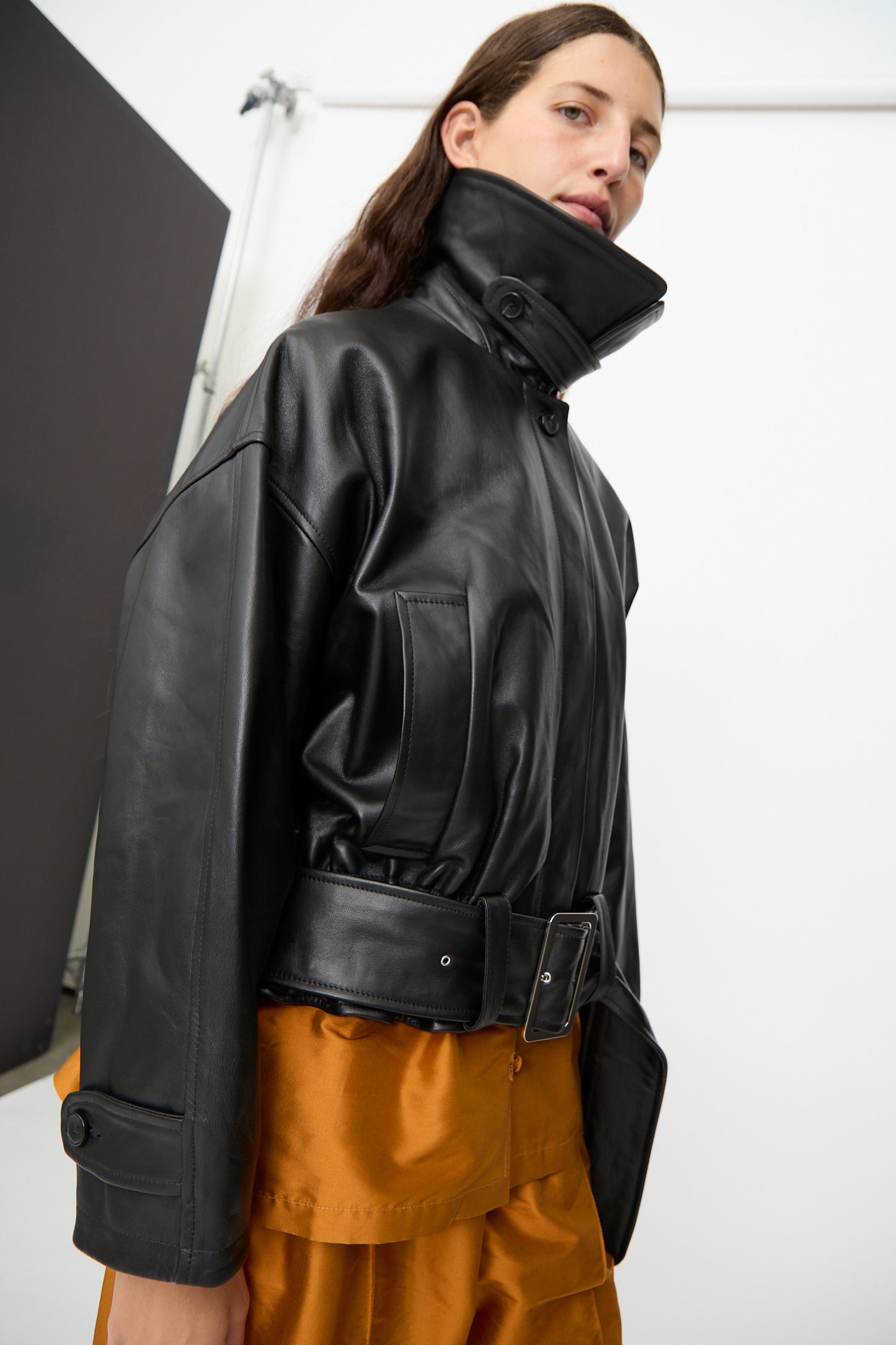 A person wearing the Cawley Nappa Leather Nara Jacket in Black, featuring a high collar and wide belt, paired with a brown skirt and standing against a plain white background.