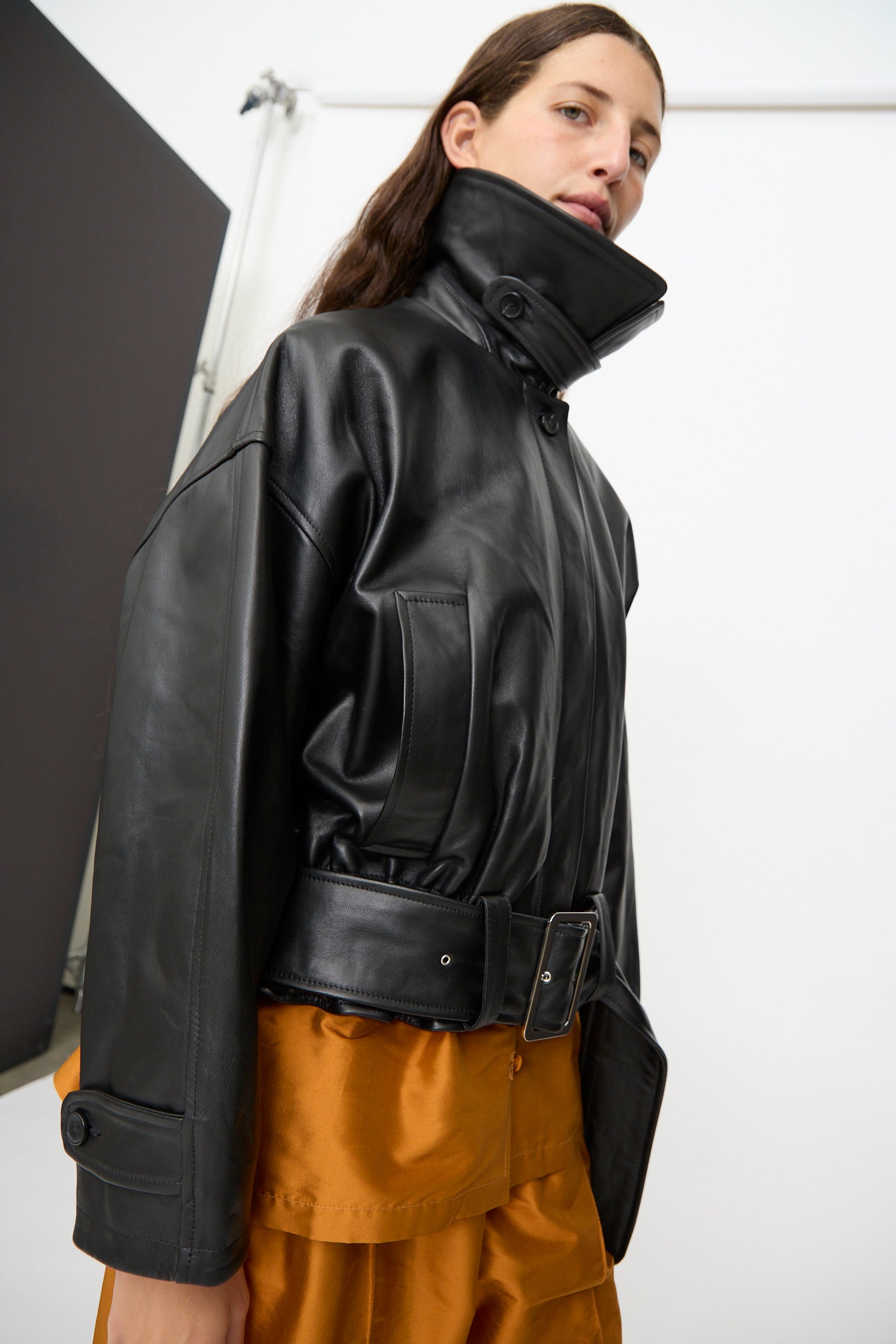 A person wearing the Cawley Nappa Leather Nara Jacket in Black, featuring a high collar and wide belt, paired with a brown skirt and standing against a plain white background.
