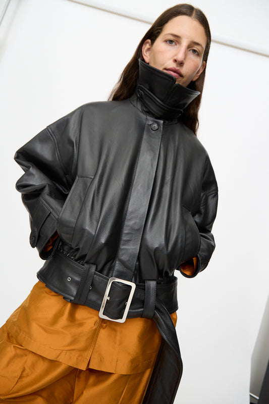 A woman wears the Cawley Nappa Leather Nara Jacket in Black—an oversized blouson style with a high collar and large buckle—paired with orange pants, standing against a plain white background.