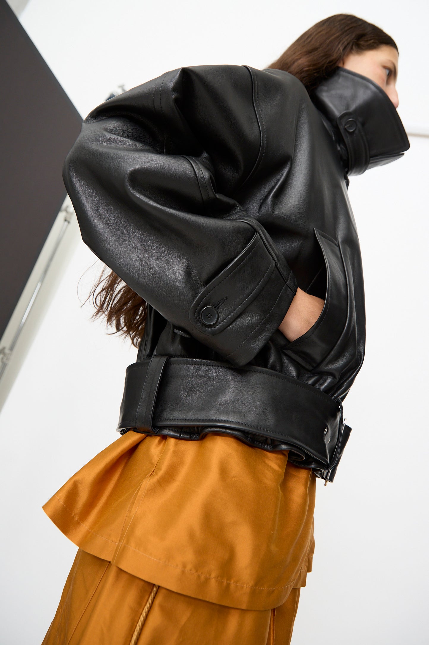 A person wearing the Cawley Nappa Leather Nara Jacket in Black—an oversized design featuring a large collar and cuffs—paired with a rust-colored skirt, stands against a plain background.