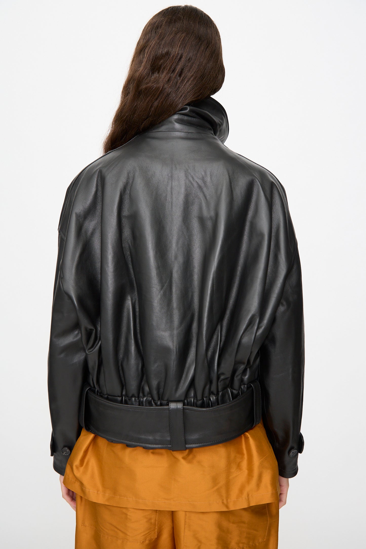 A person with long brown hair is photographed from the back against a plain white background, wearing Cawley’s oversized Nappa Leather Nara Jacket in black paired with orange pants.
