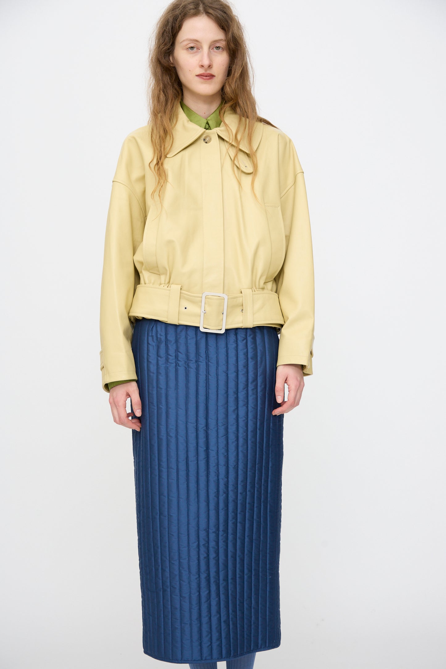 A person stands facing forward wearing the Cawley Nappa Leather Nara Jacket in Butter, made in London, over a green shirt and a long quilted blue skirt.