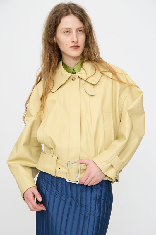 A person with long wavy hair wears the Cawley Nappa Leather Nara Jacket in Butter over a green shirt and blue quilted skirt, standing against a plain white background.