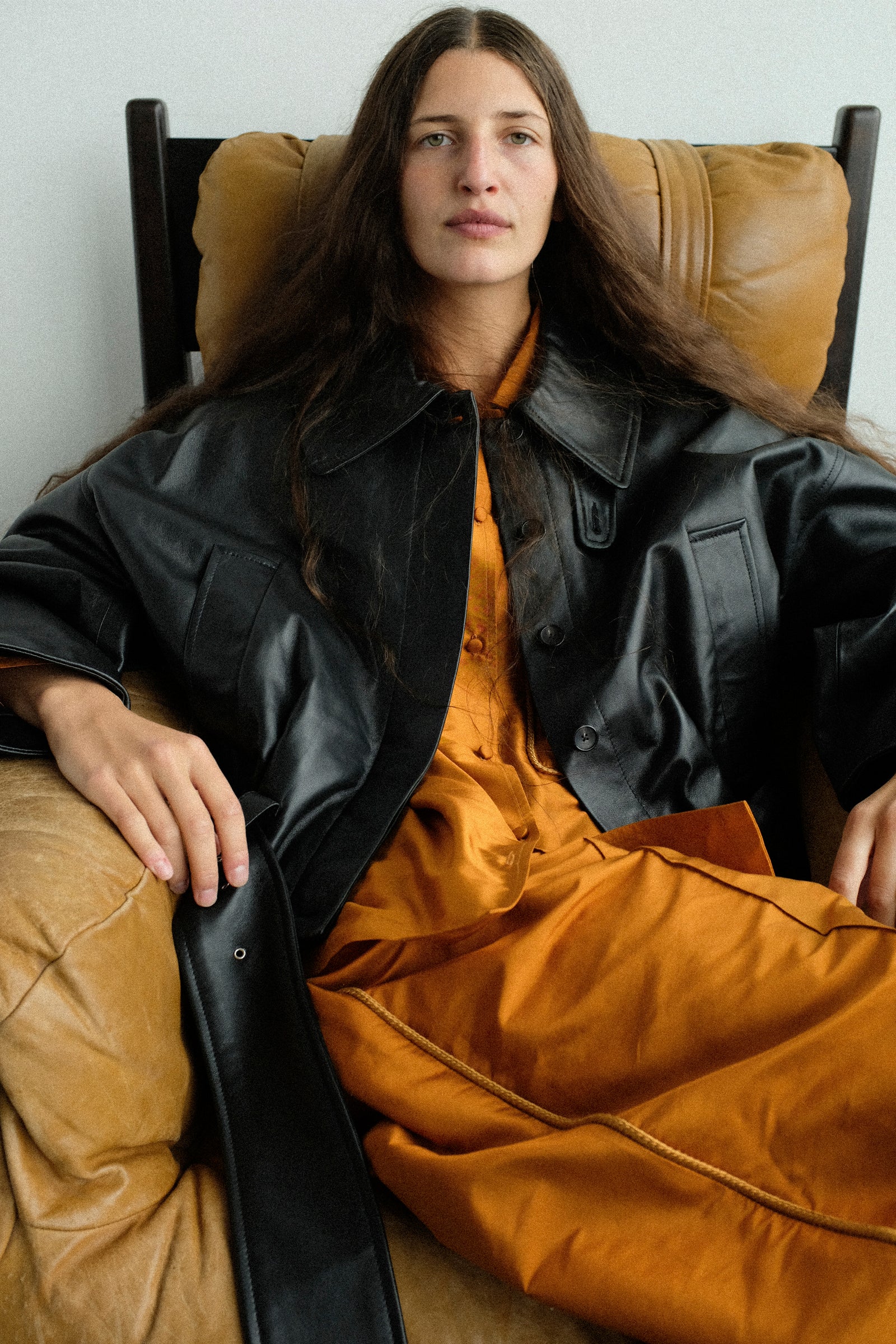 A woman reclining in a tan leather armchair, wearing Cawley's Silk Dupion August Shirt in Sienna, and Nappa Leather Nara Jacket in Black over.