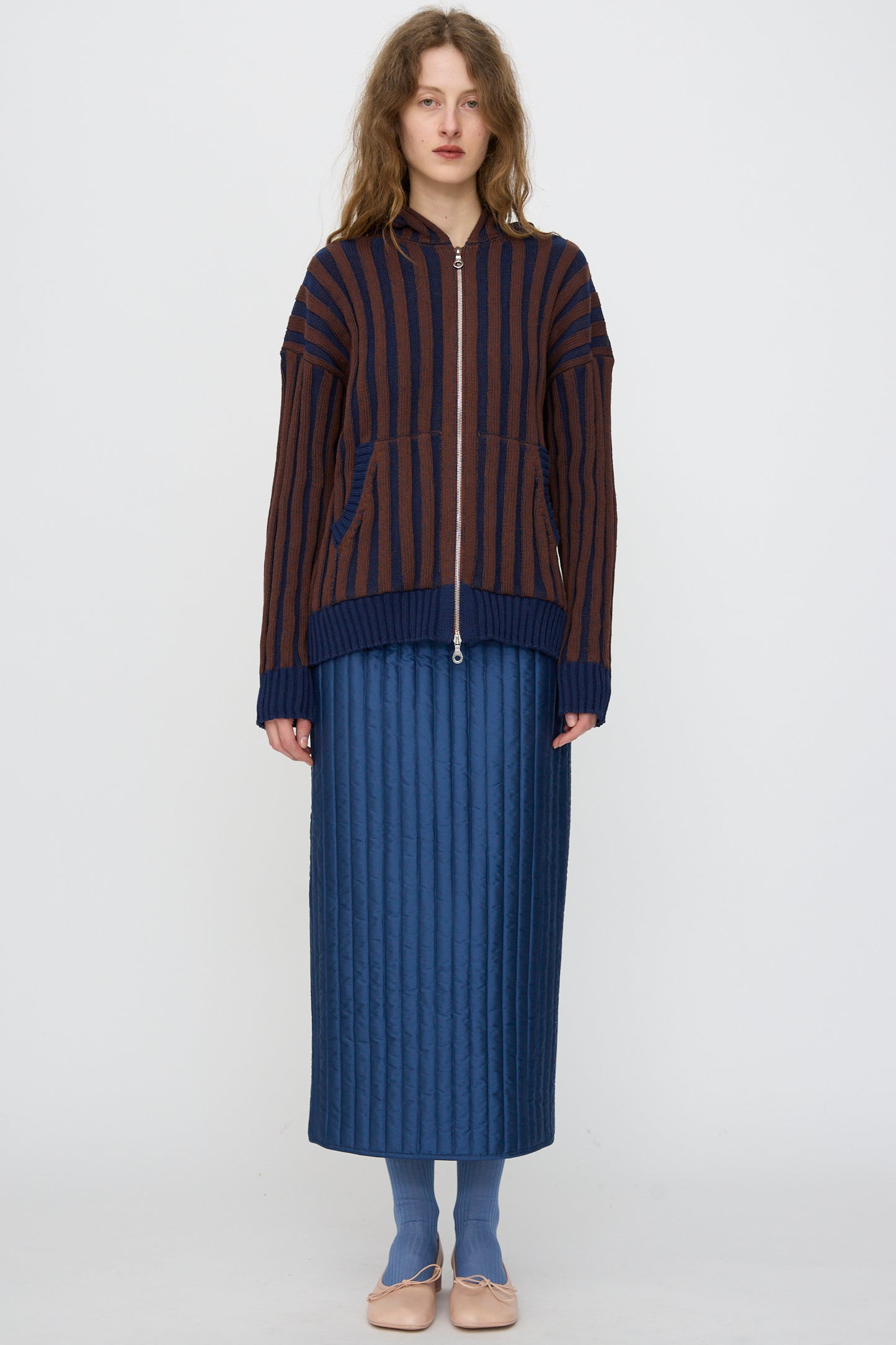 A person stands against a plain background, wearing Cawley's Organic Cotton Zip Up Hoodie in Blu Kid and Choco, styled with a quilted blue skirt, blue socks, and beige flats.