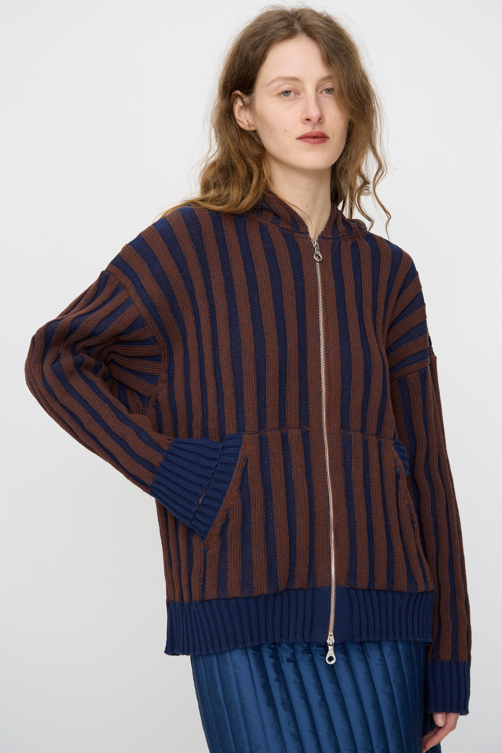 A person wears the Cawley Organic Cotton Zip Up Hoodie in Blu Kid and Choco, featuring ribbed stripes and front pockets, paired with a blue quilted skirt, standing against a plain background.
