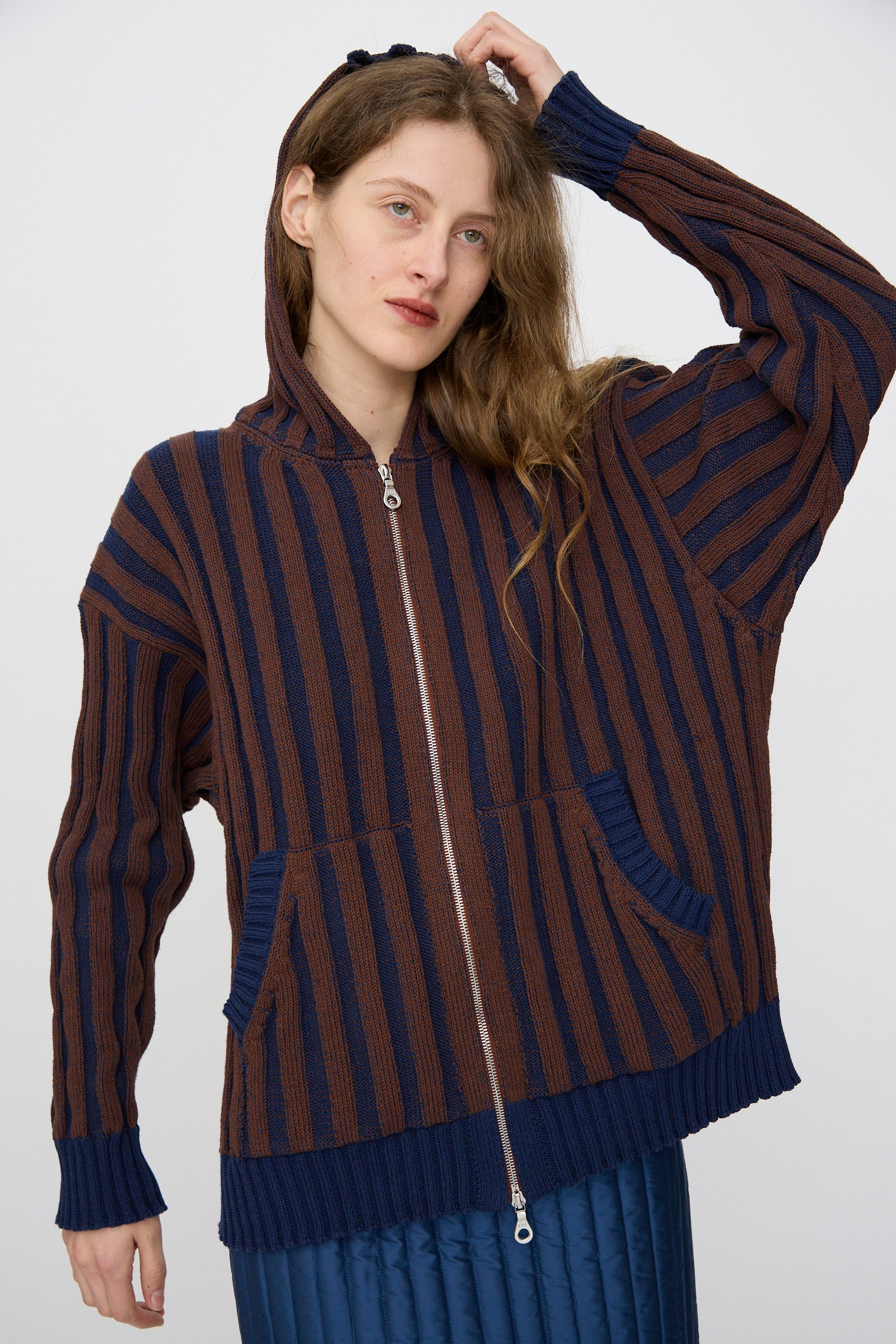 A person wearing the Cawley Organic Cotton Zip Up Hoodie in Blu Kid and Choco, styled with front pockets and a relaxed fit, paired with a blue quilted skirt and standing with one hand on their head.