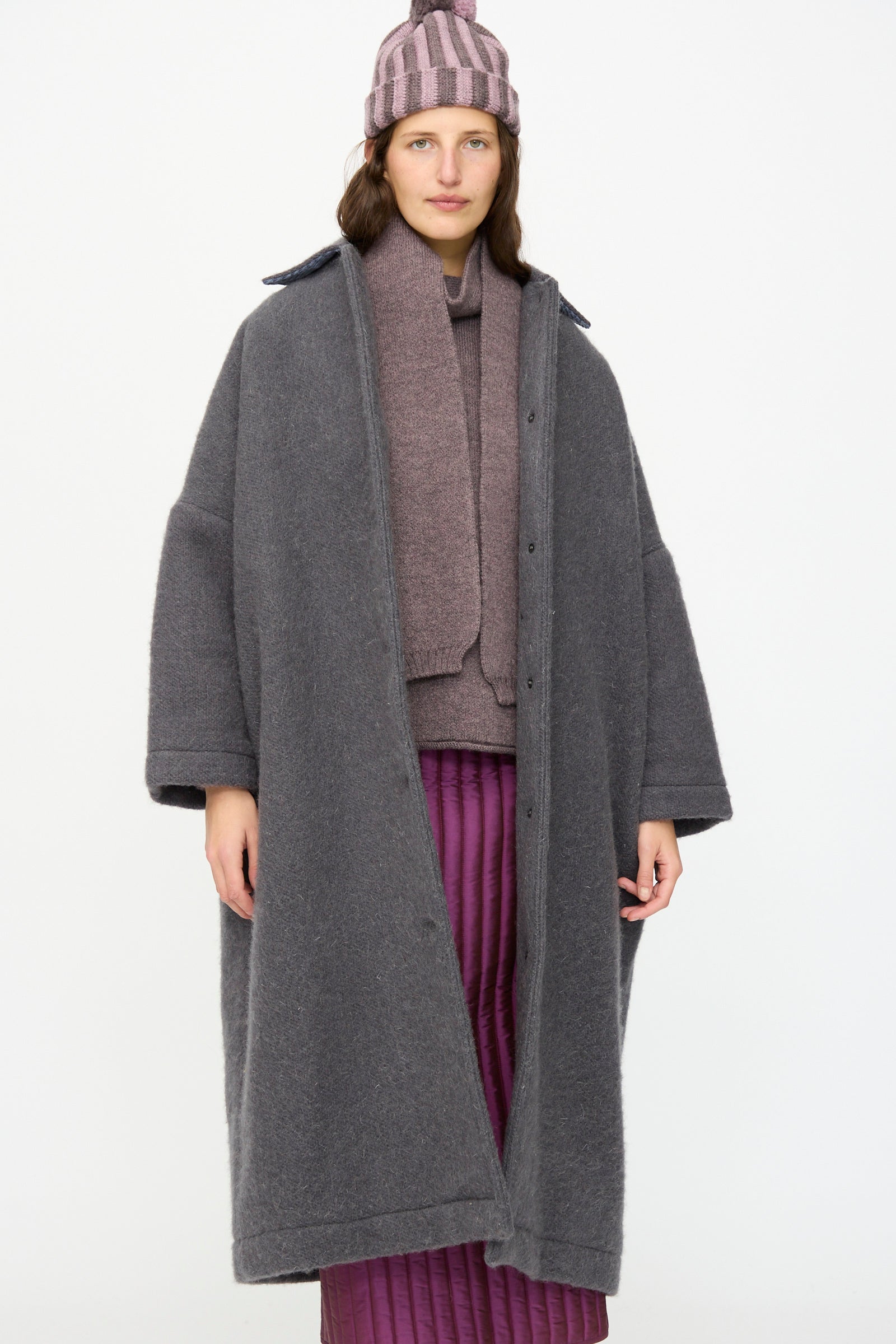 A person wears the Cawley Portuguese Hairy Wool Faro Coat in Slate, paired with a mauve sweater, scarf, striped pink beanie, and a quilted purple skirt, standing against a plain white background.