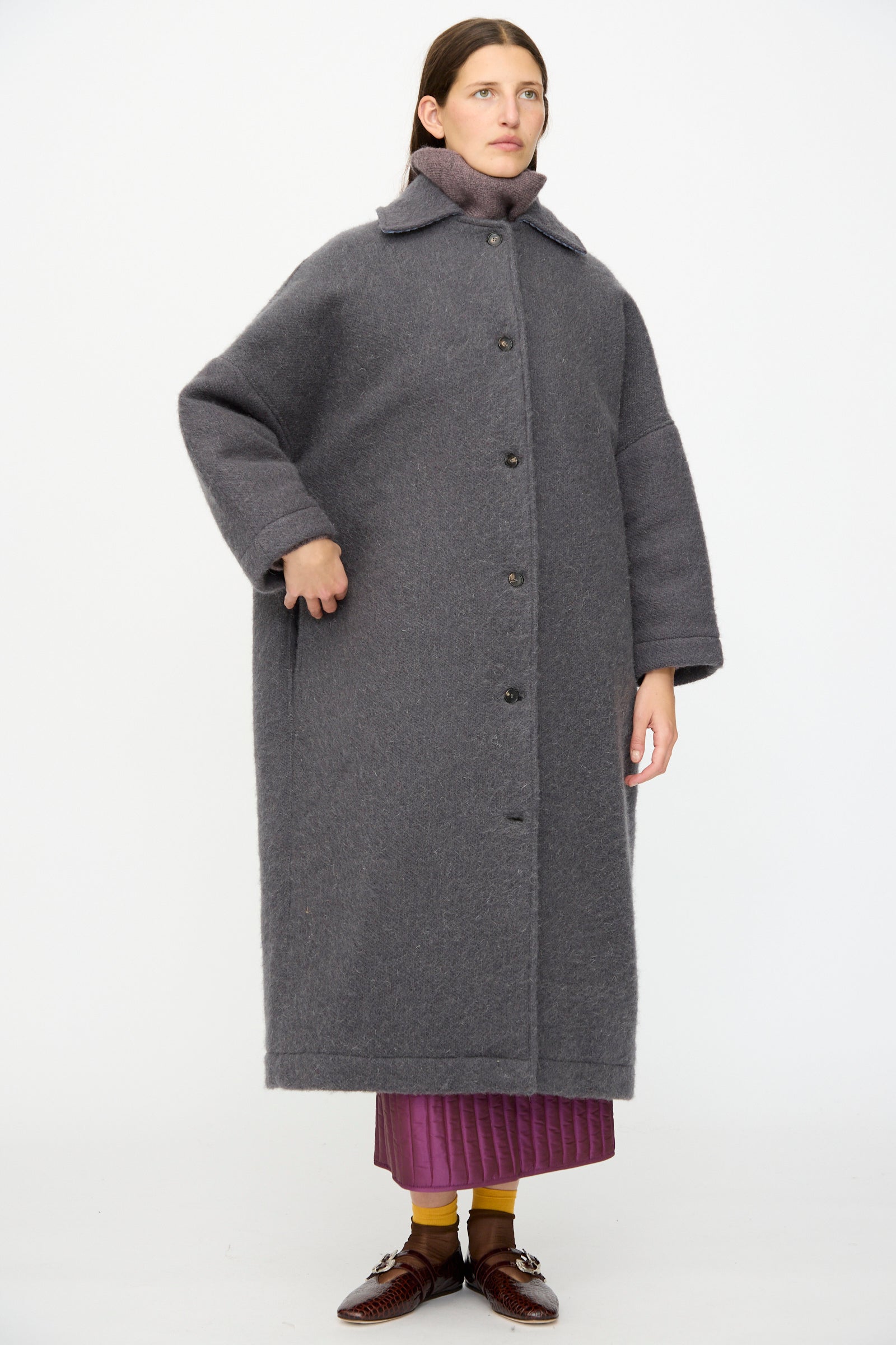 A person stands against a white background wearing the Cawley Portuguese Hairy Wool Faro Coat in Slate, buttoned over a pink skirt, paired with brown shoes and yellow socks.