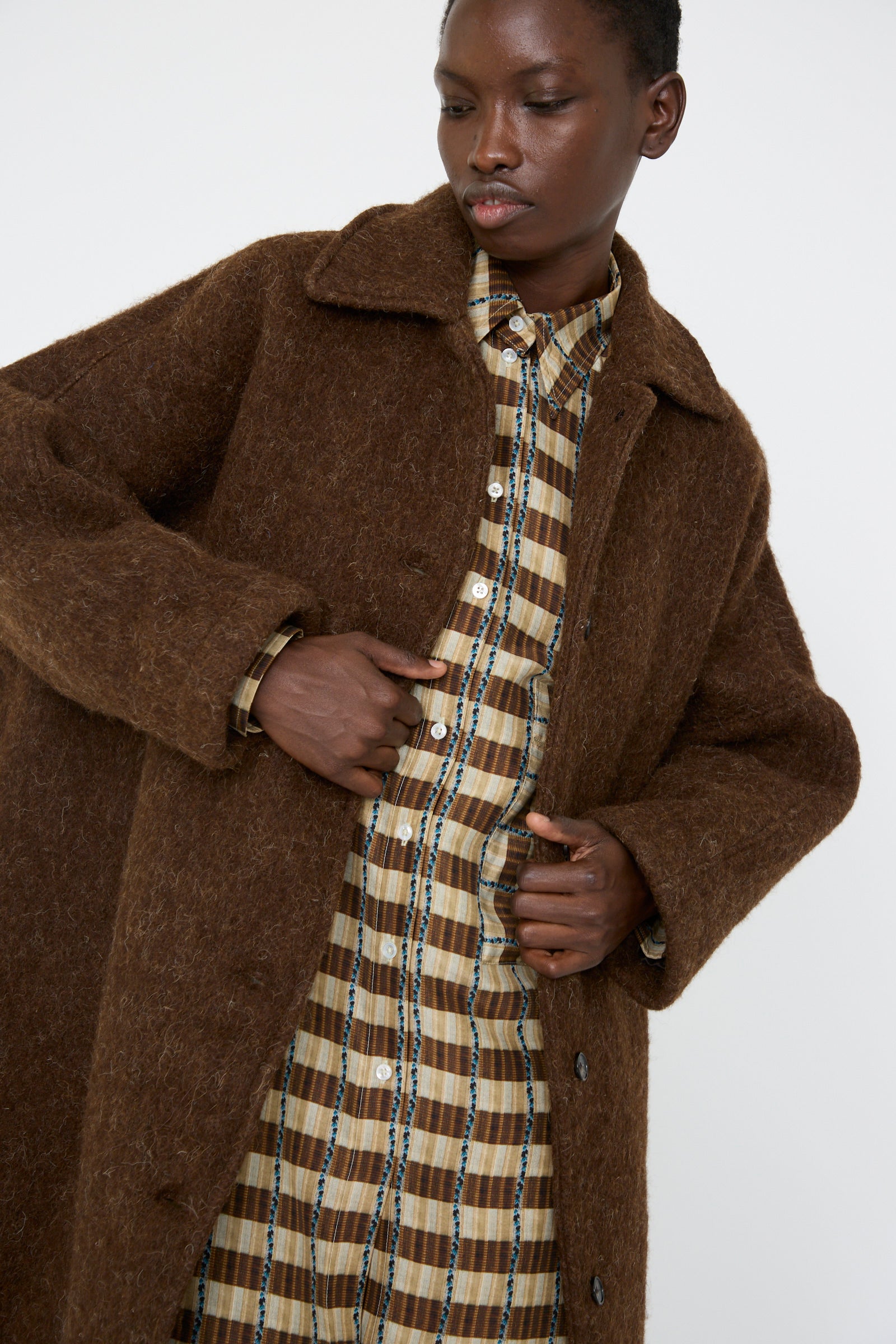 A person wears the Portuguese Long Hair Wool Faro Coat in Chocolate by Cawley over a plaid dress, set against a plain background.