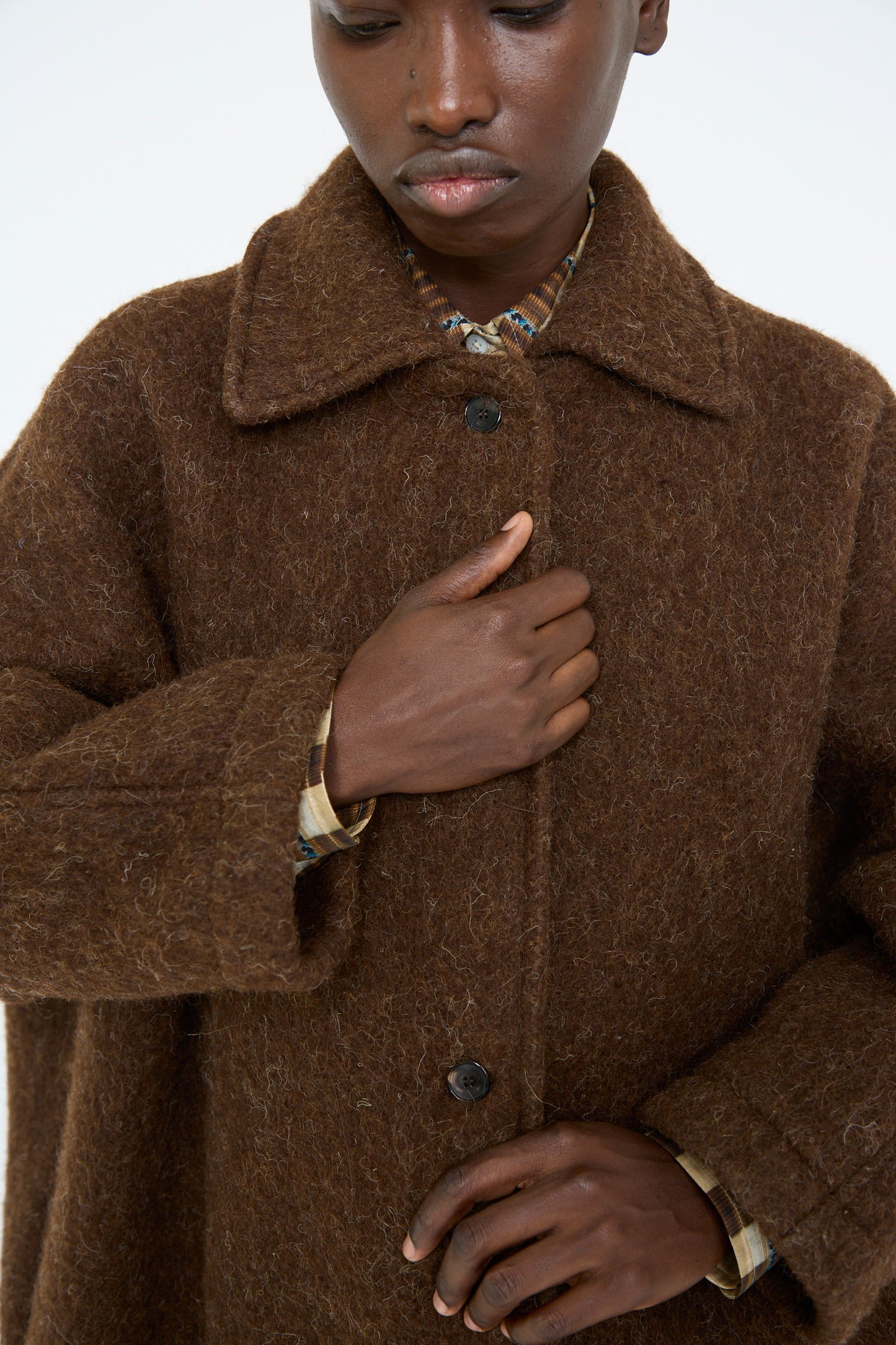 A person stands against a plain background and looks down, wearing the Cawley Portuguese Long Hair Wool Faro Coat in Chocolate, which features an oversized fit.