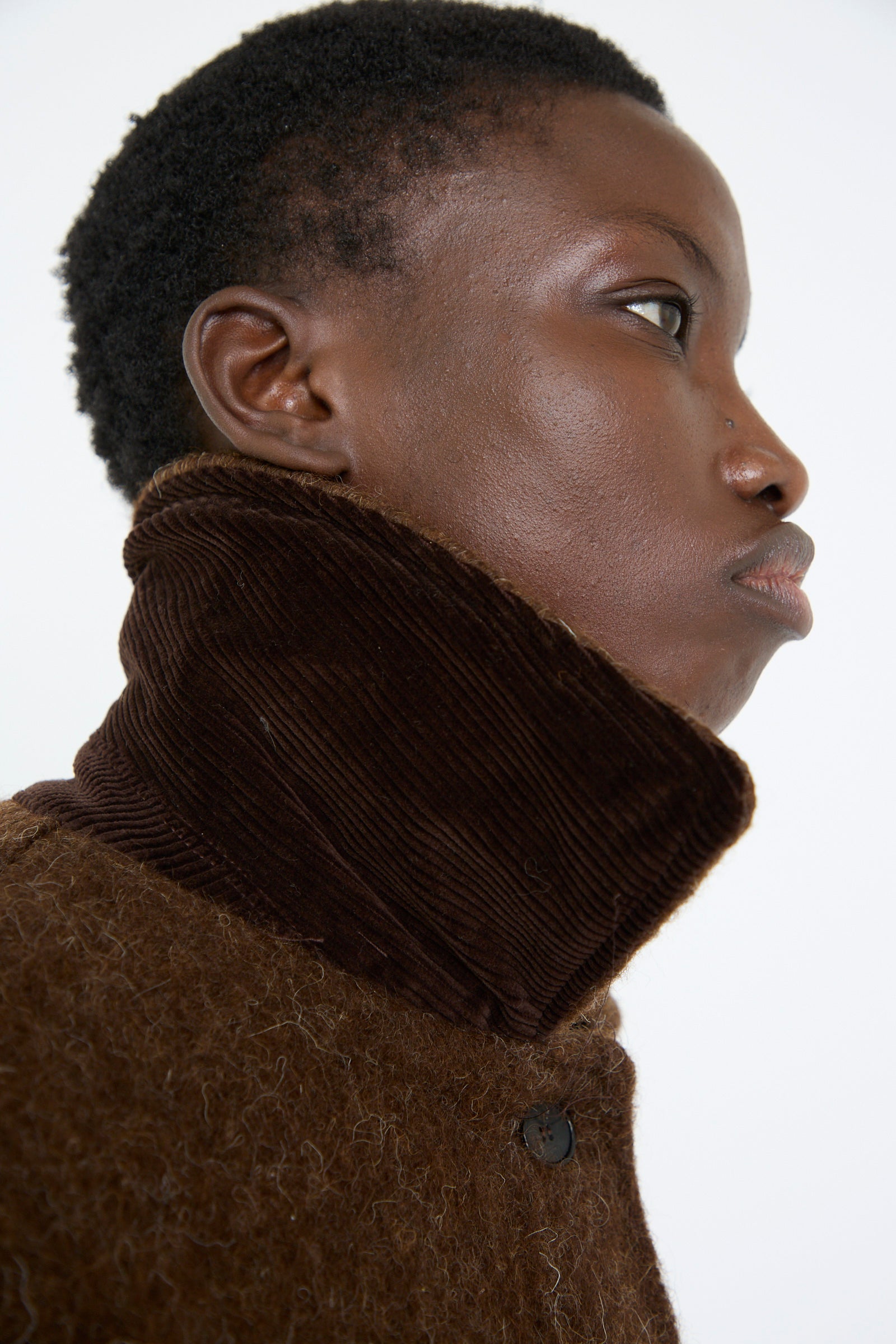 A close up of a woman wearing a Cawley Portuguese Long Hair Wool Faro Coat in Chocolate, looking to the side against a plain background.