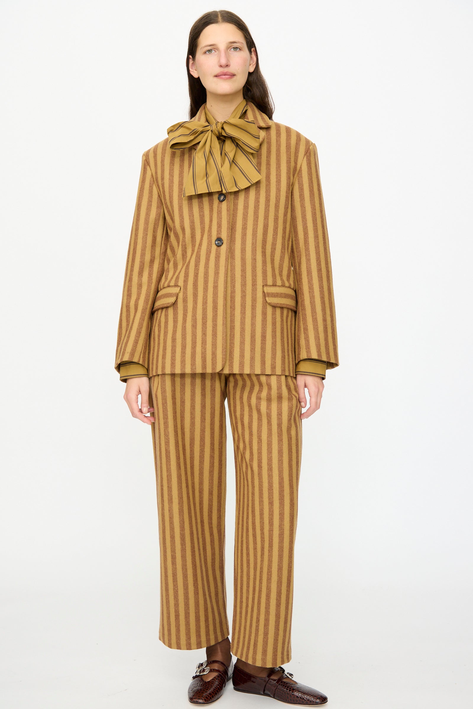 A person stands against a plain background wearing Cawley’s Portuguese Striped Wool Andie Blazer in Goldfinch and Bordeaux, styled with wide legs, a matching neck bow, and brown shoes—an effortlessly chic look made in London.