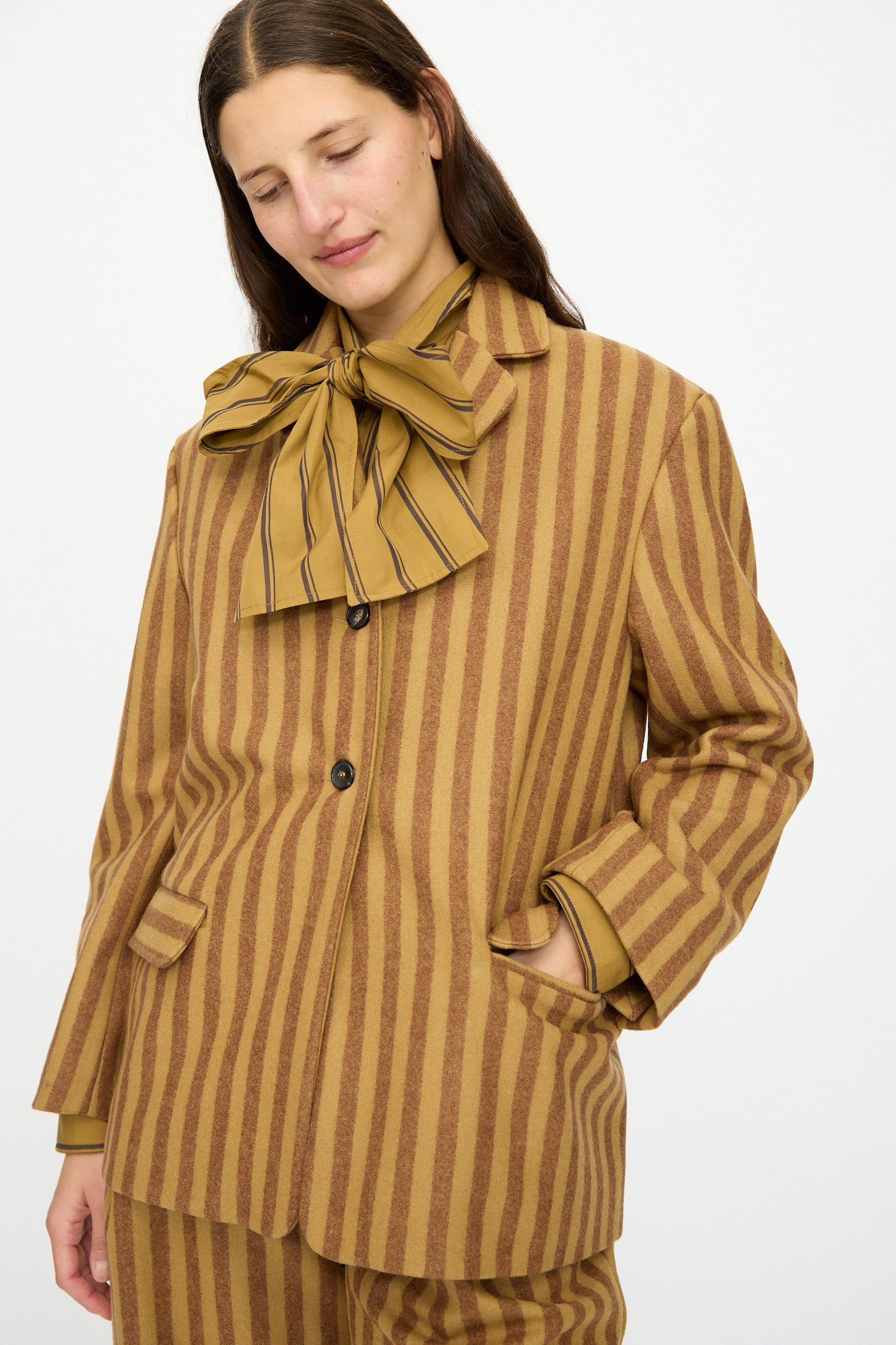 A person wearing the Cawley Portuguese Striped Wool Andie Blazer in Goldfinch and Bordeaux, featuring brown and tan vertical stripes with a large matching bow, stands with one hand in their pocket and looks down. Made in London.