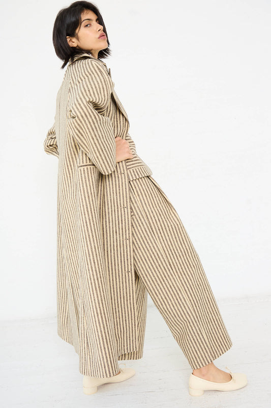 A person with short dark hair stands sideways, wearing the Cawley Portuguese Striped Wool Annie Coat in Ecru and Brown and matching wide-legged pants. The outfit is beige with horizontal stripes, and the relaxed fit is complemented by light-colored shoes.