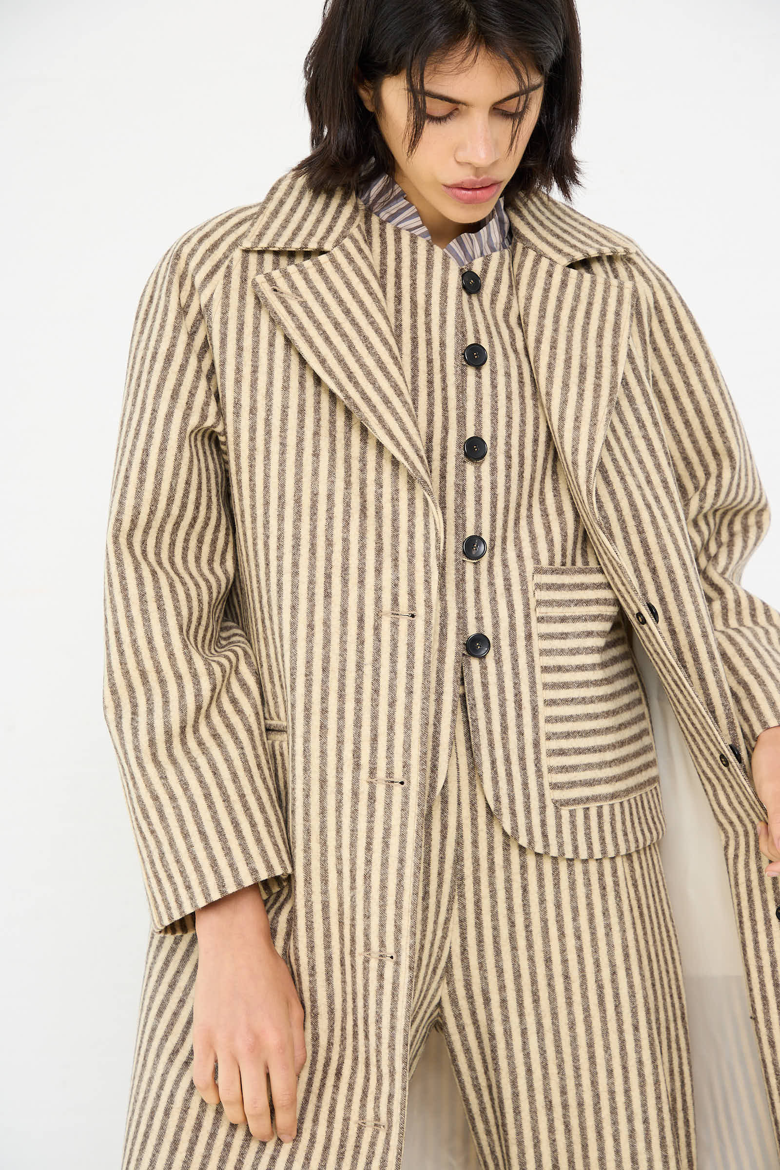 A person wearing the Cawley Portuguese Striped Wool Annie Coat in Ecru and Brown over a matching outfit, adjusting the coat with one hand.