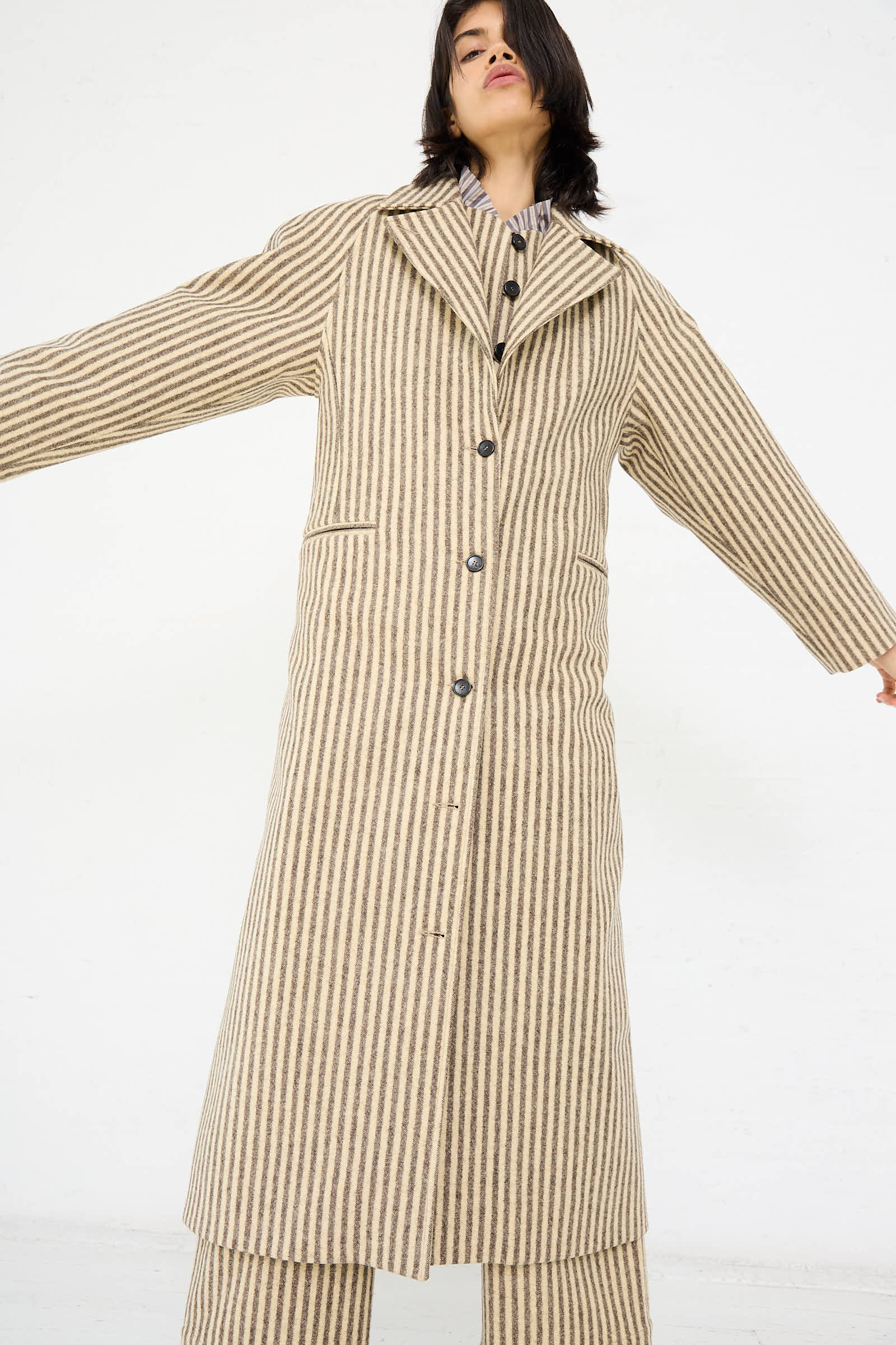 Person wearing the Portuguese Striped Wool Annie Coat in Ecru and Brown by Cawley with outstretched arms, posing against a plain white background.