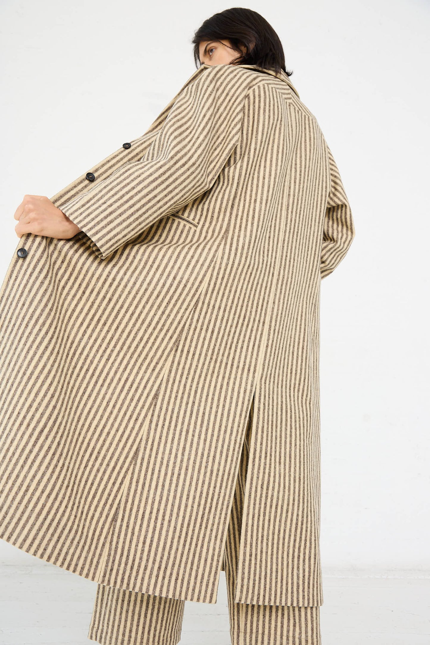 A person wearing the Cawley Portuguese Striped Wool Annie Coat in Ecru and Brown with a relaxed fit, standing with their back slightly turned to showcase the coat's design and buttons on the sleeves.