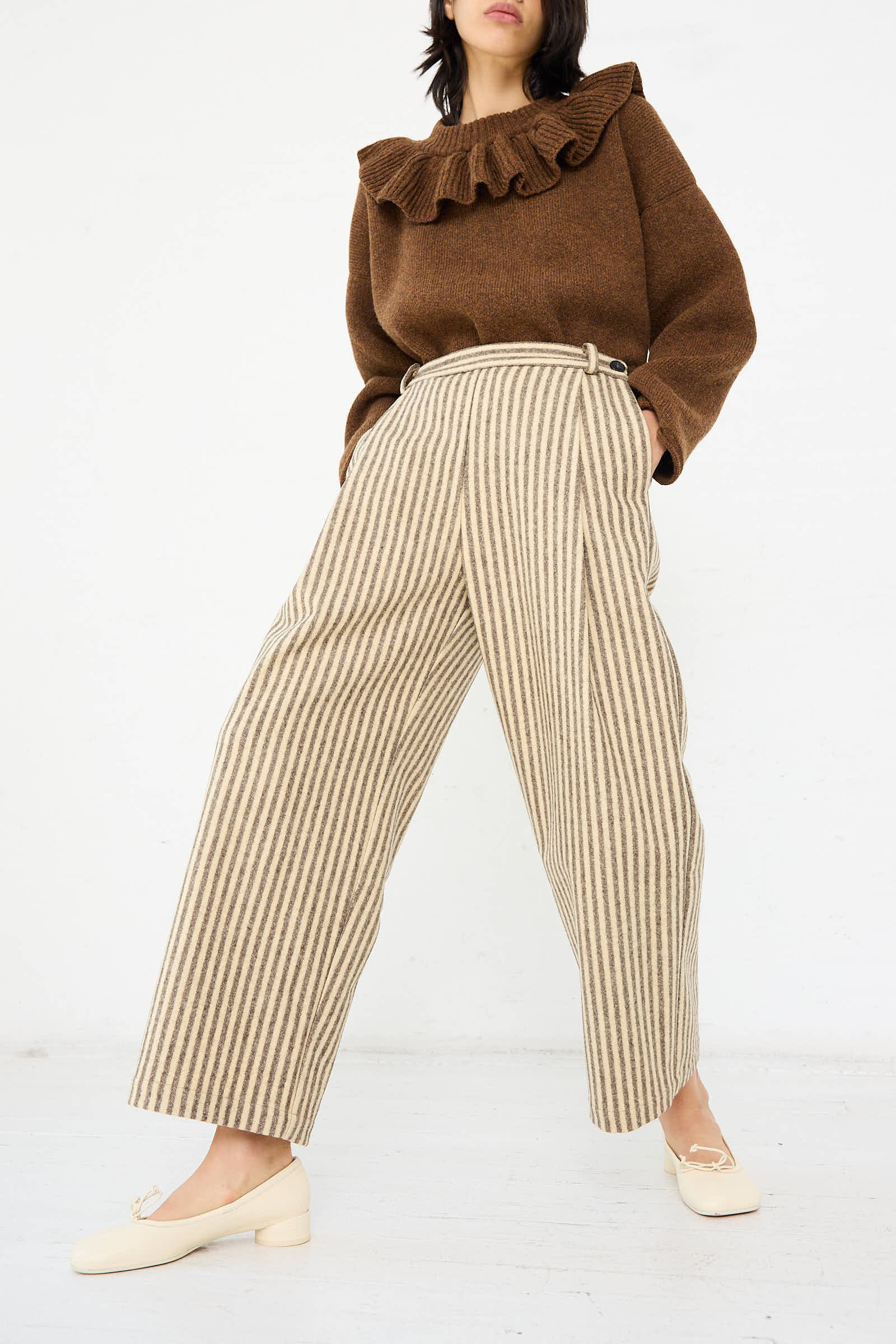 A person is wearing a brown ruffled sweater, Portuguese Striped Wool Georgia Trousers in Ecru and Brown by Cawley, and off-white loafers. They are standing against a plain white background.