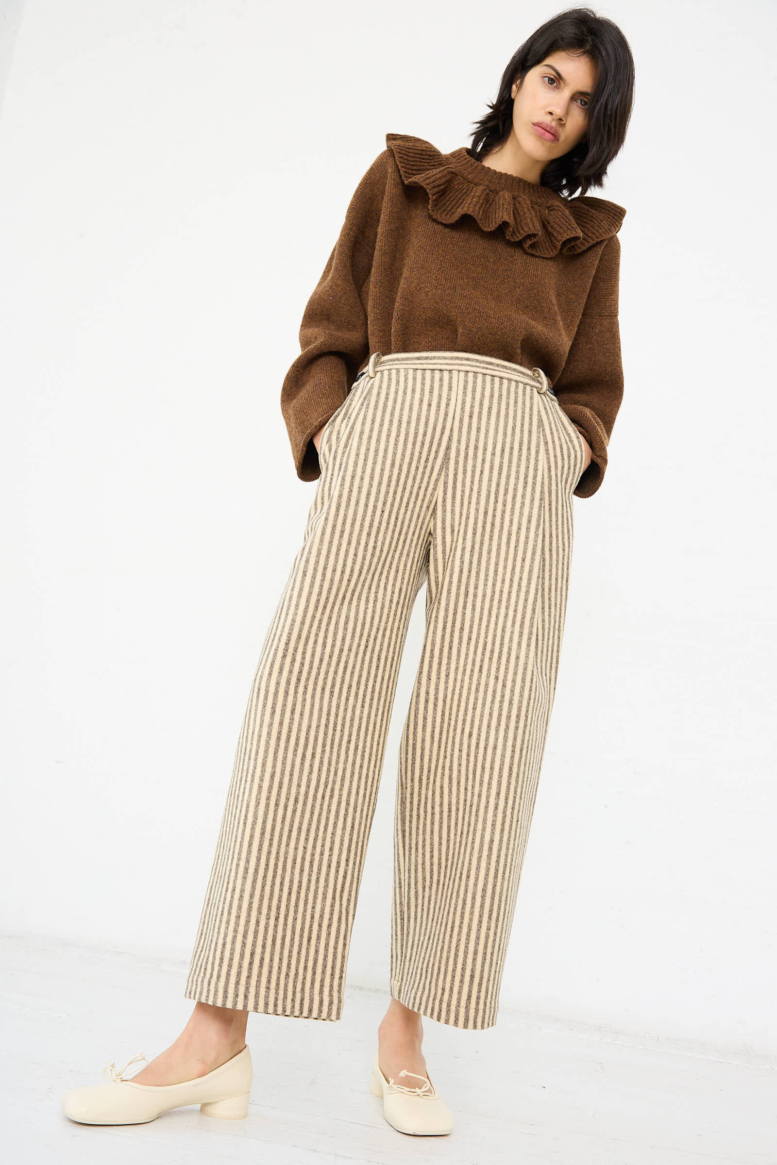 Person standing against a plain background, wearing a brown ruffled sweater, Cawley's Portuguese Striped Wool Georgia Trousers in Ecru and Brown with a relaxed fit, and white shoes.