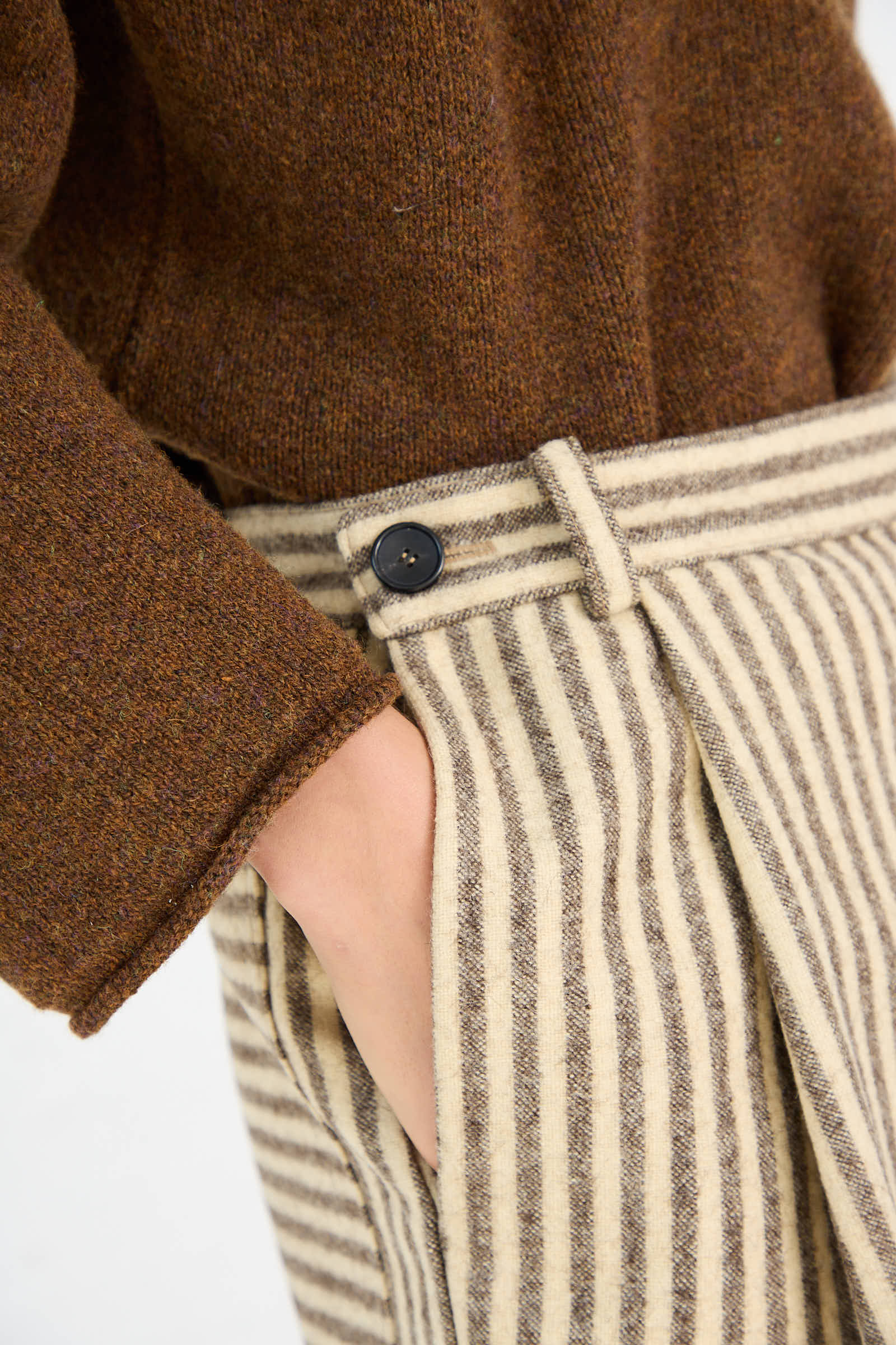 Close-up of a person's hand in the pocket of relaxed fit, high-rise Portuguese Striped Wool Georgia Trouser in Ecru and Brown by Cawley, paired with a brown wool sweater. The pants feature a buttoned waistband.