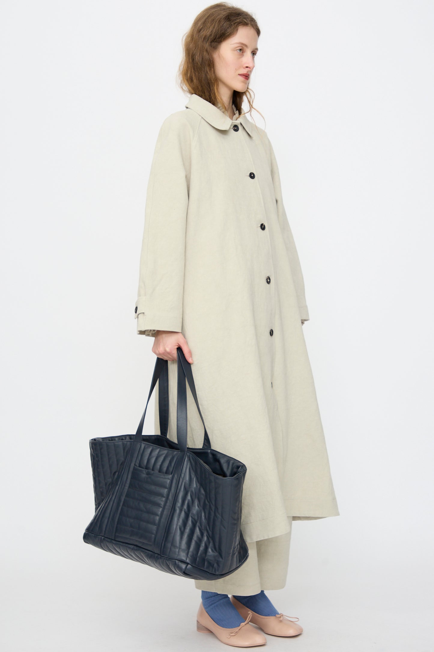 A person holds the Cawley Quilted Nappa Large Tote Bag with Coin Purse in Navy against a plain white background while wearing a long, light-colored coat and beige shoes.