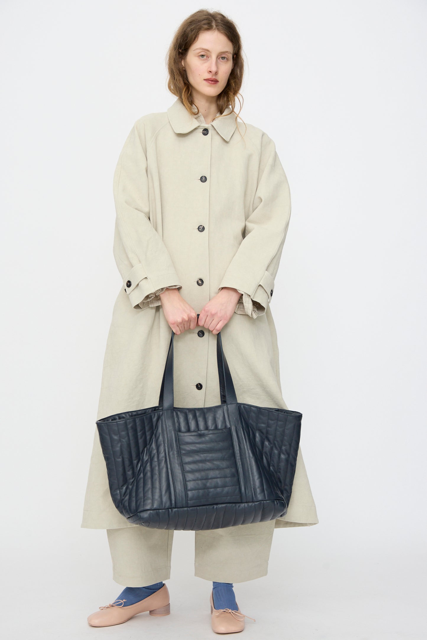 A woman in a long beige coat and pants holds the Cawley Quilted Nappa Large Tote Bag with Coin Purse in Navy, standing against a plain white background.