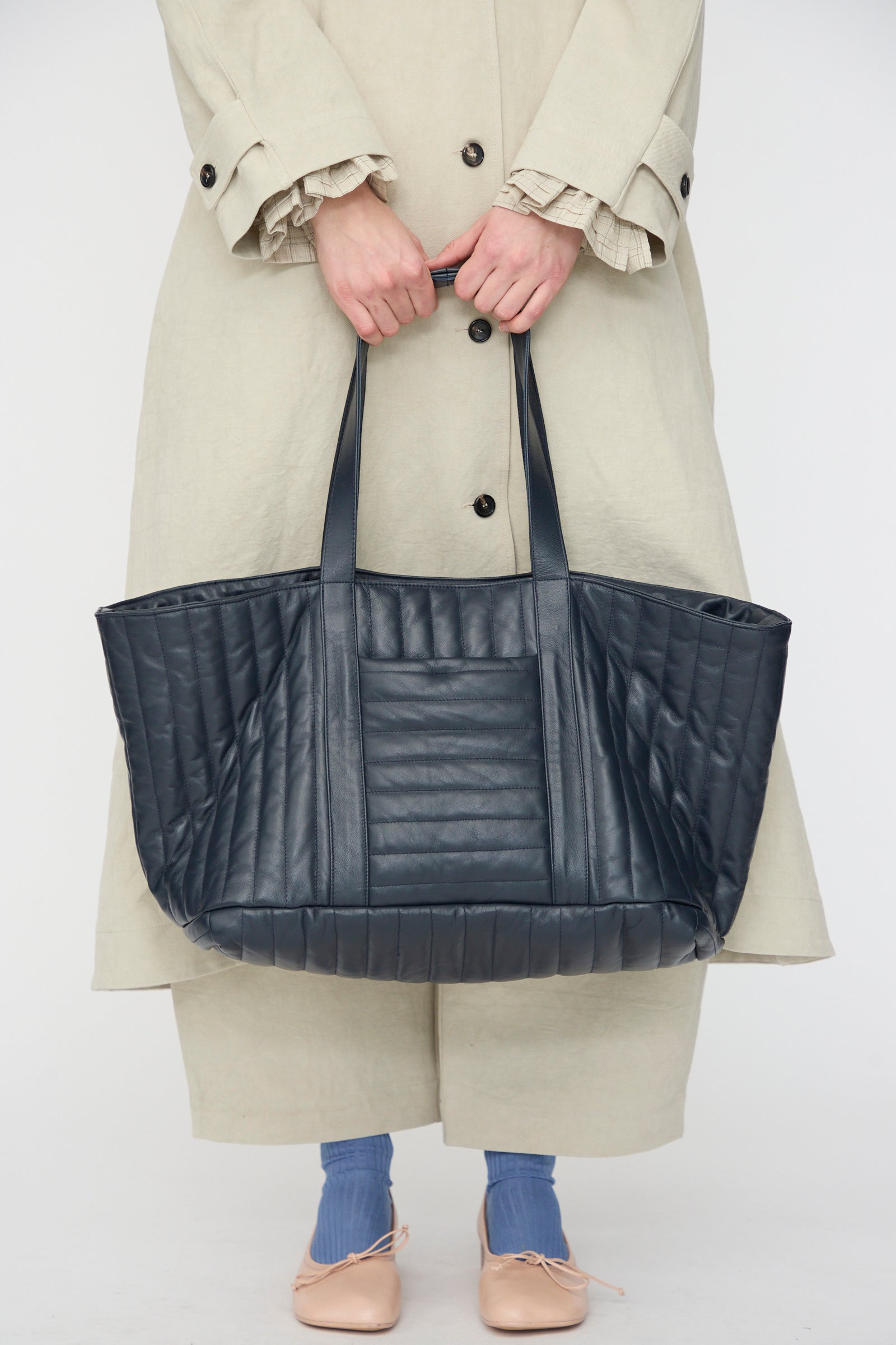 A person in a beige coat holds the Cawley Quilted Nappa Large Tote Bag with Coin Purse in Navy. Visible are only blue socks, beige shoes, handcrafted in London.