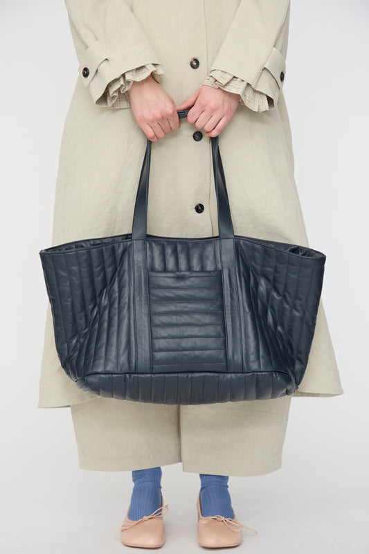 A person in a beige coat holds the Cawley Quilted Nappa Large Tote Bag with Coin Purse in Navy. Visible are only blue socks, beige shoes, handcrafted in London.