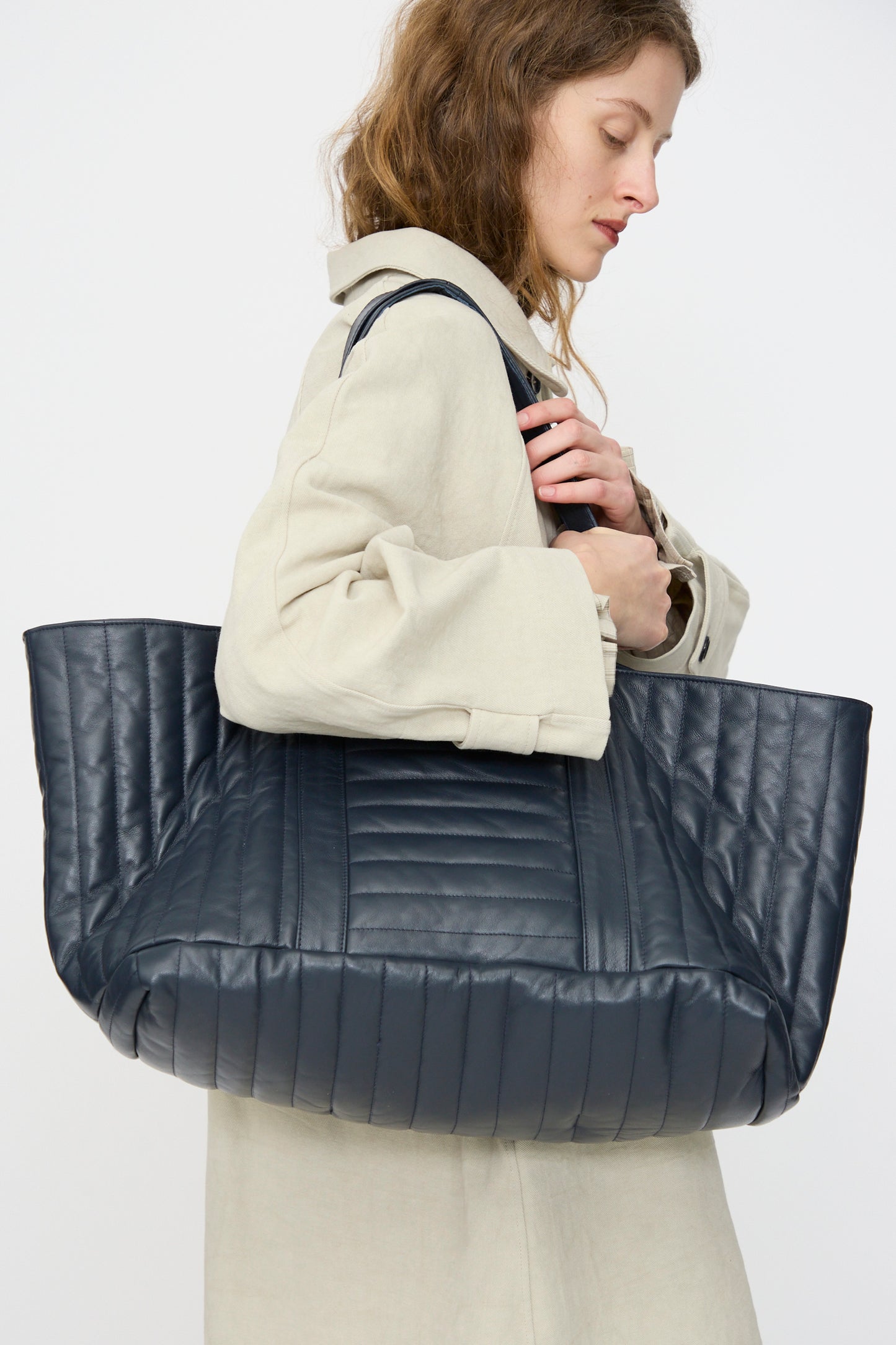 A person in a beige coat holds the Cawley Quilted Nappa Large Tote Bag with Coin Purse in Navy over their shoulder, standing against a plain background.