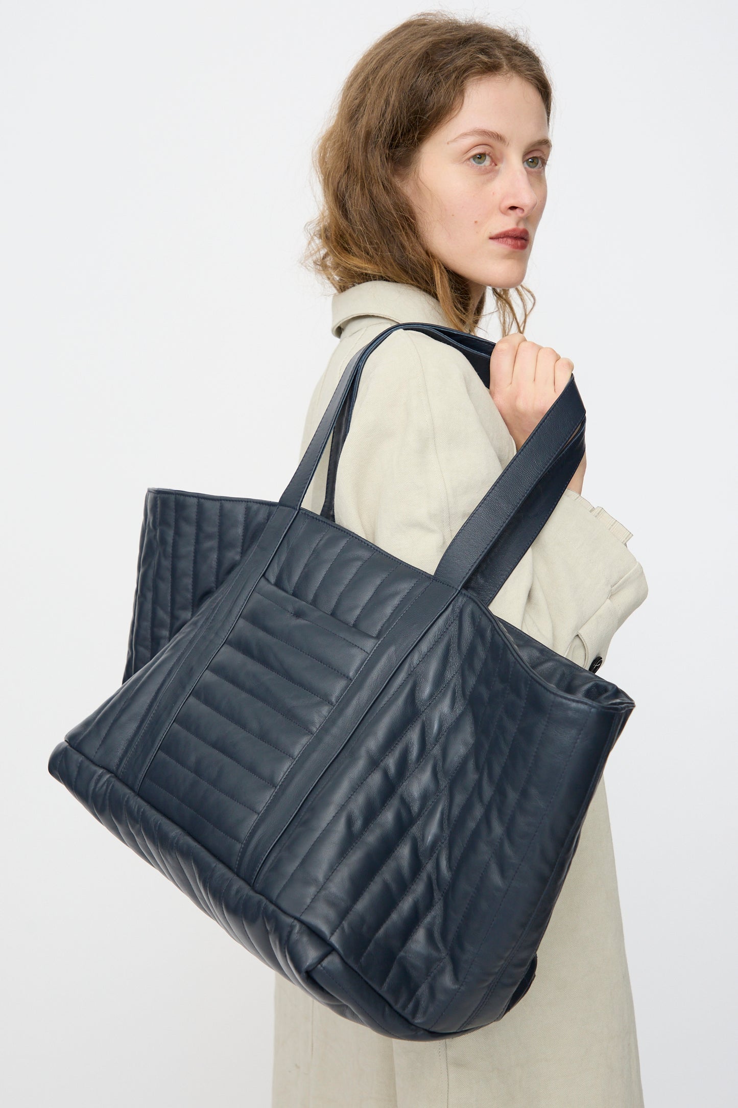 A woman in a light coat holds the Cawley Quilted Nappa Large Tote Bag with Coin Purse in Navy over her shoulder, looking slightly to the side against a plain background.