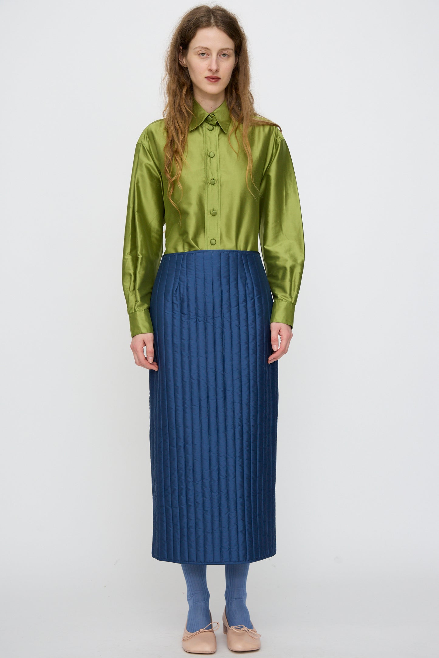 A woman stands against a plain background wearing a shiny green button-up shirt, the Cawley Quilted Silk Sadie Skirt in Insignia, blue socks, and beige flats.